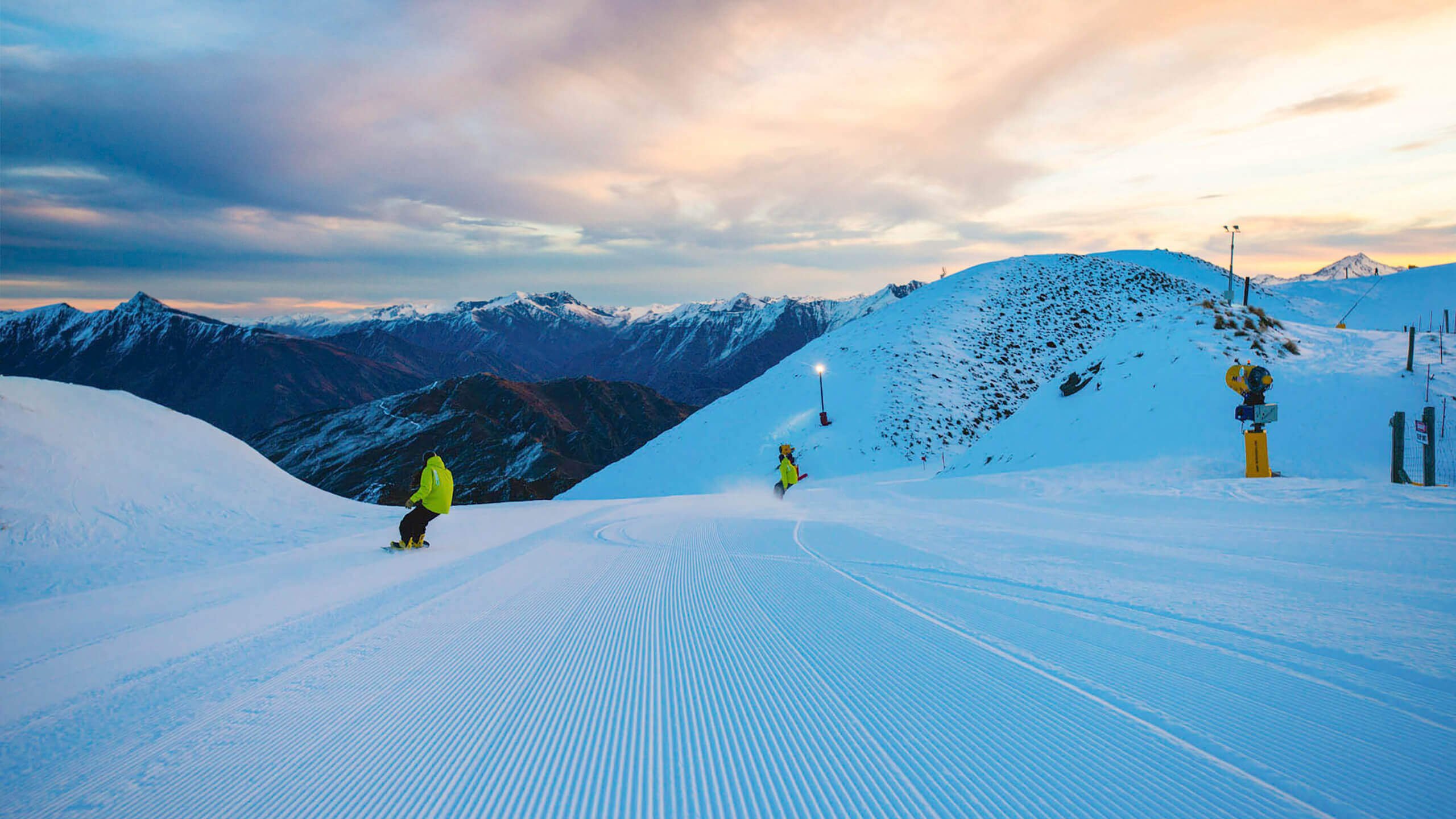 Two snowboarders riding on freshly groomed slopes as the sun rises.
