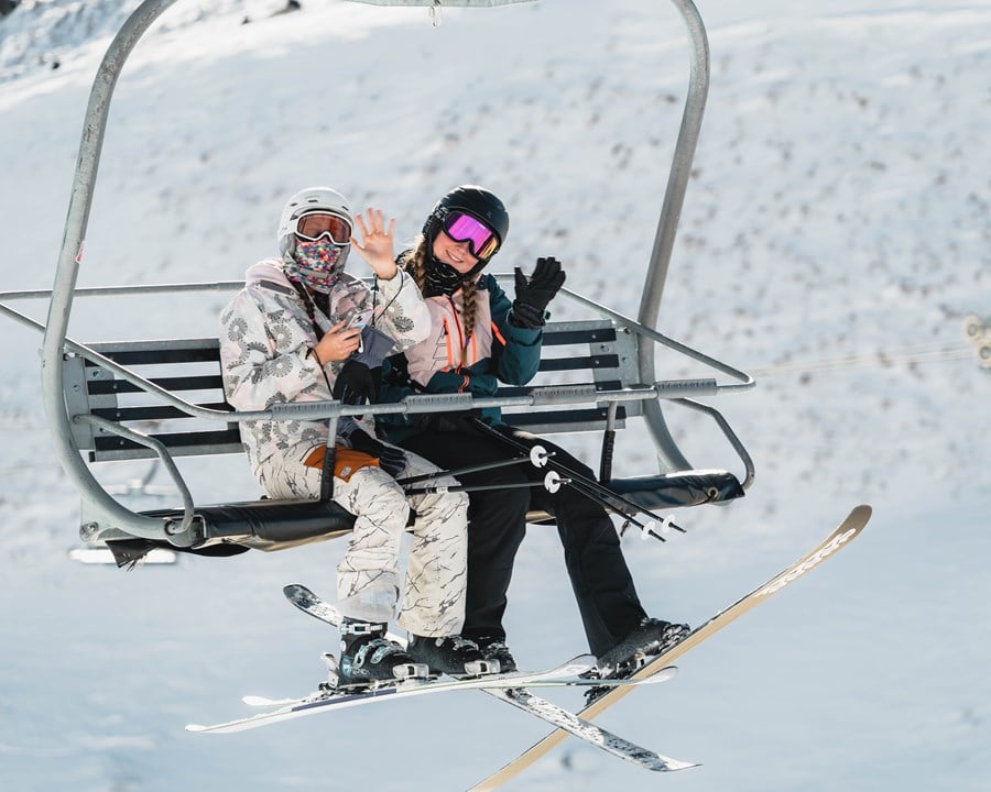 Two women sitting on a chairlift smiling and waving.