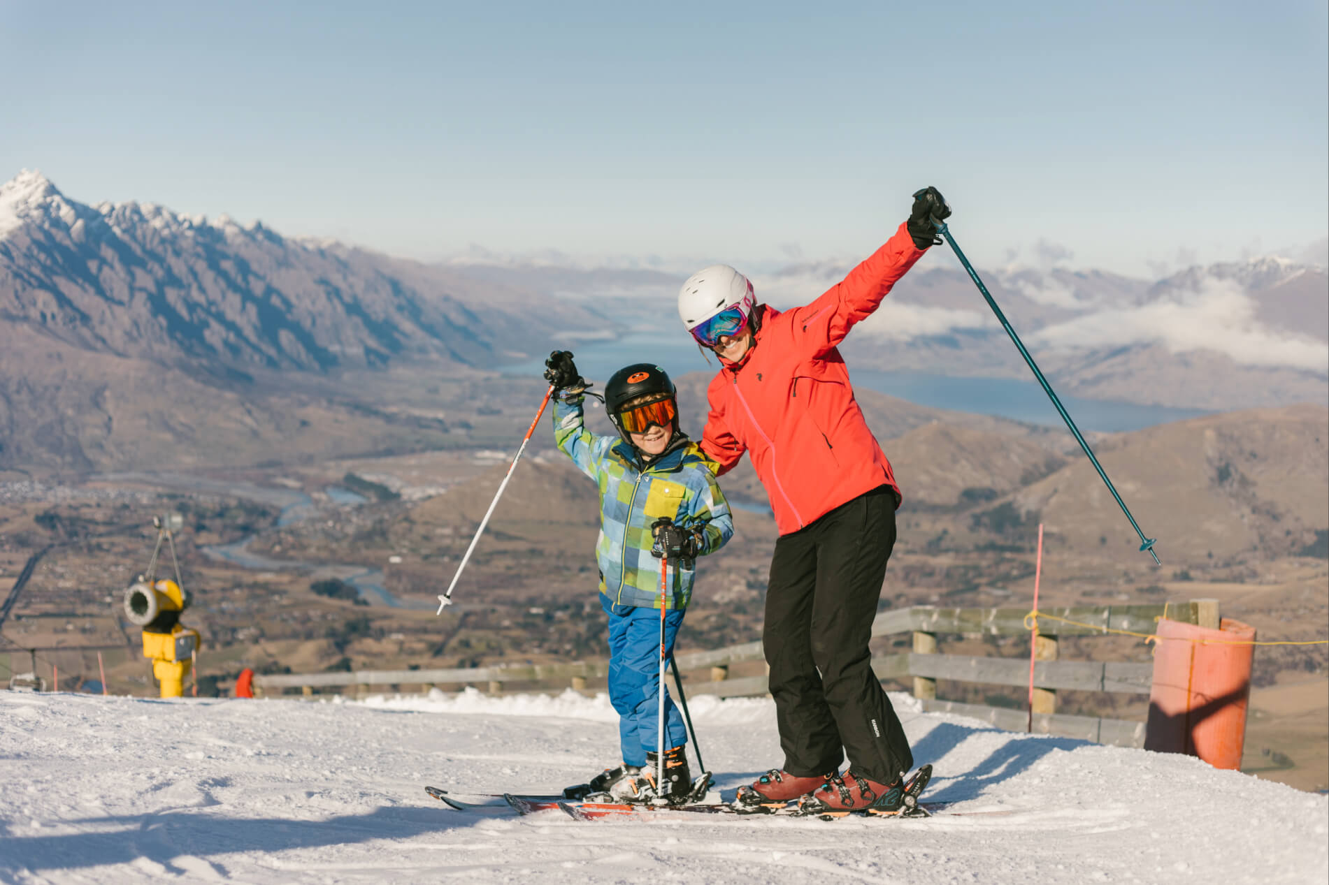 Adult and child in ski gear posing cheerfully with poles on snowy slope with mountains in the background.