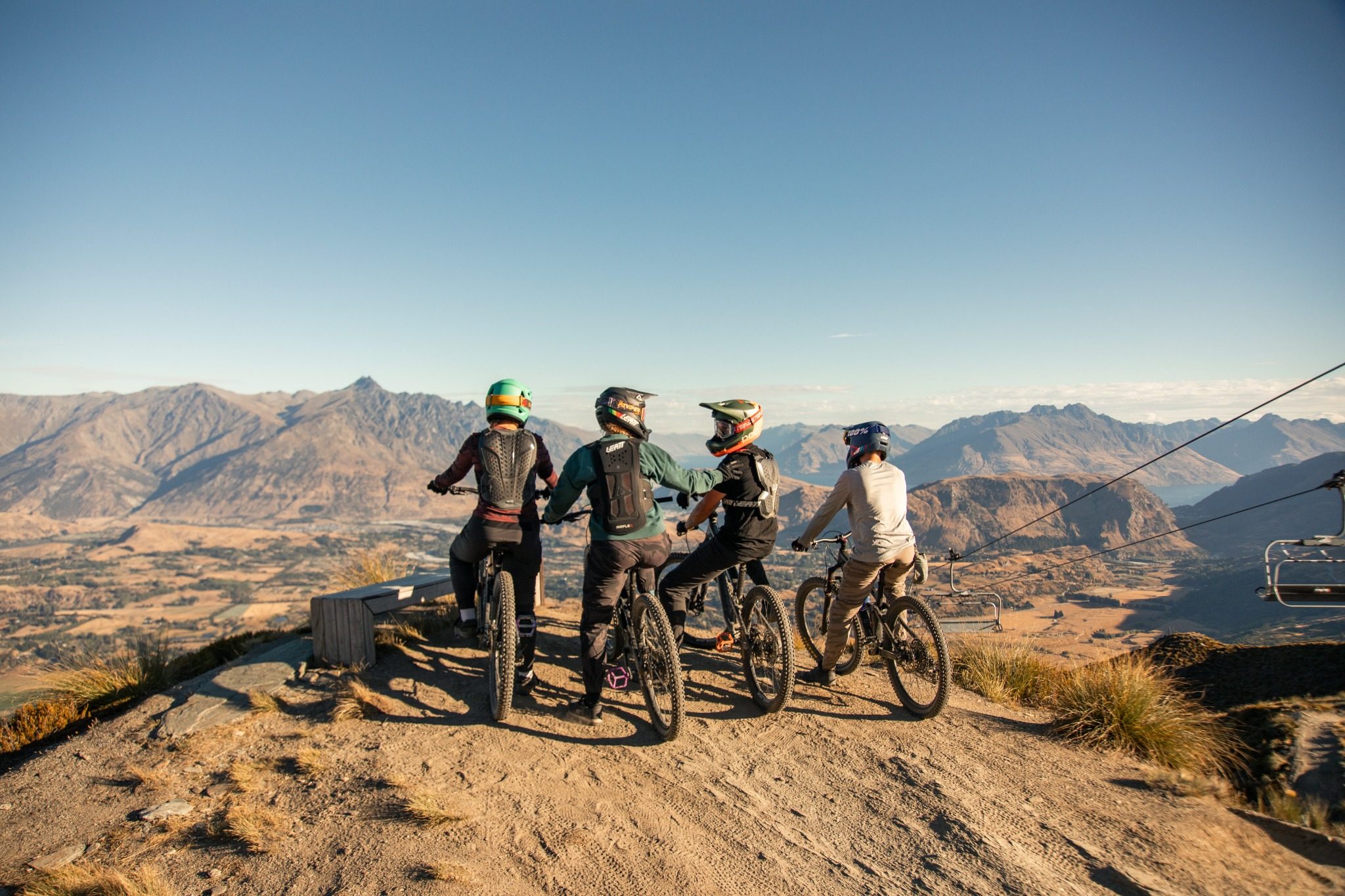 Four mountain bikers with helmets and gear face a scenic mountain valley under clear sky.