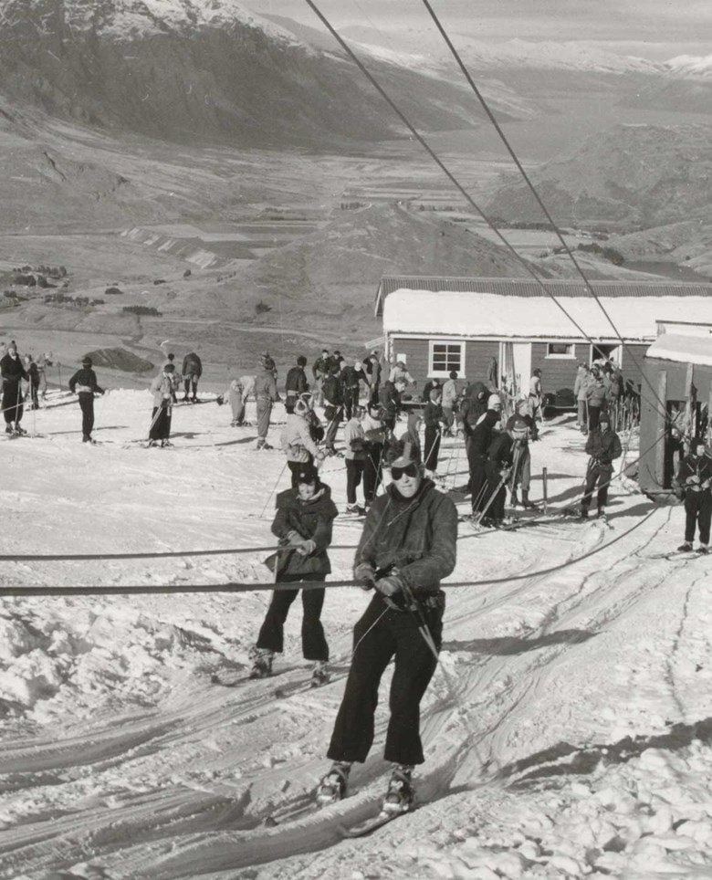 Vintage black and white photo of skiers using a ski tow at a snowy mountain resort with a lodge.