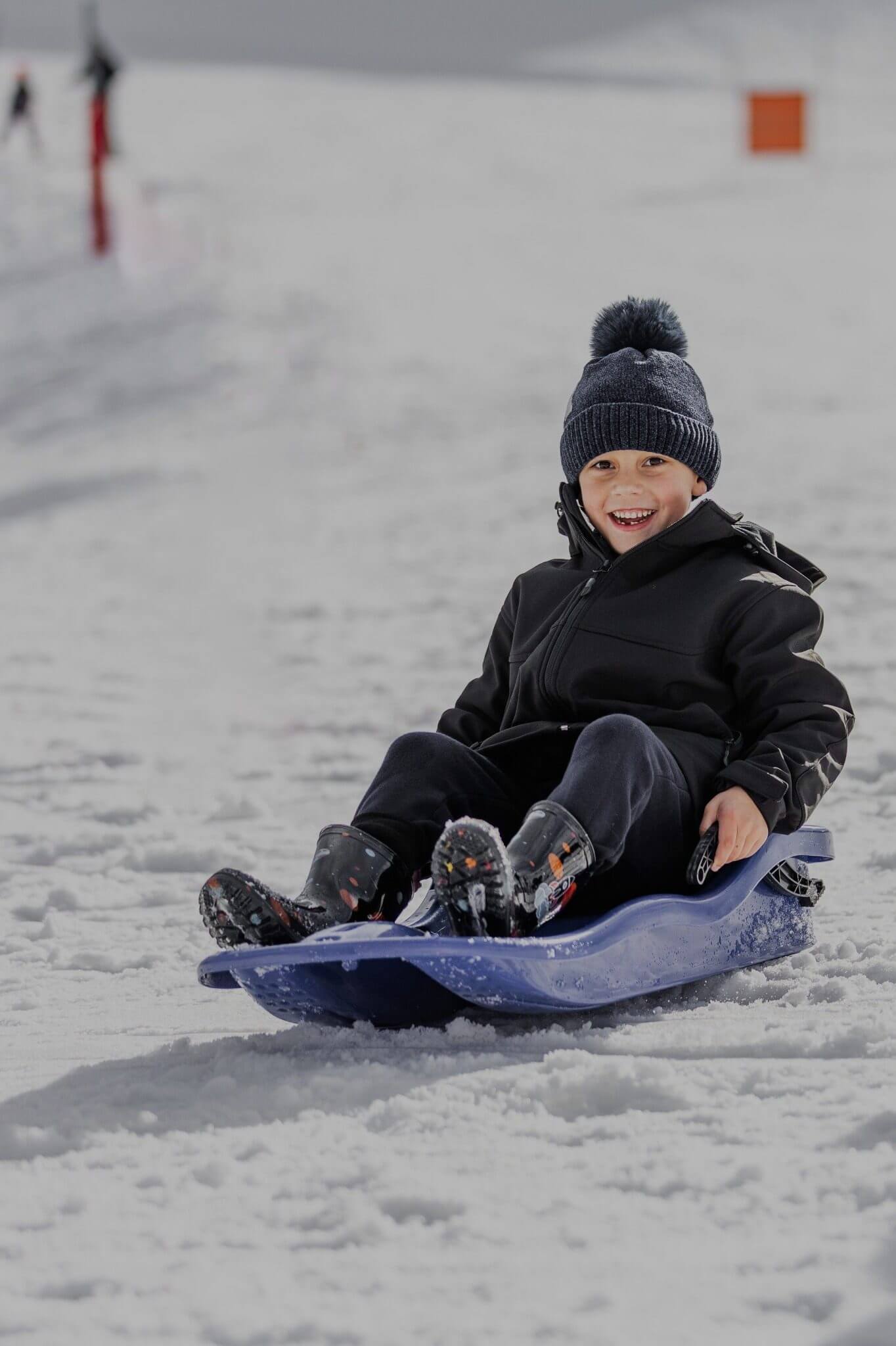 Smiling child in winter clothes sledding down a snowy hill on a blue sled.