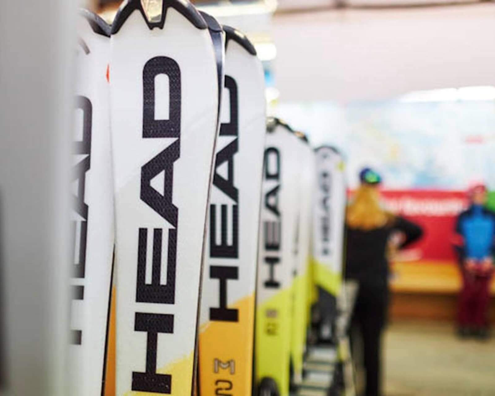 Row of HEAD brand skis standing upright indoors with blurred people in background.