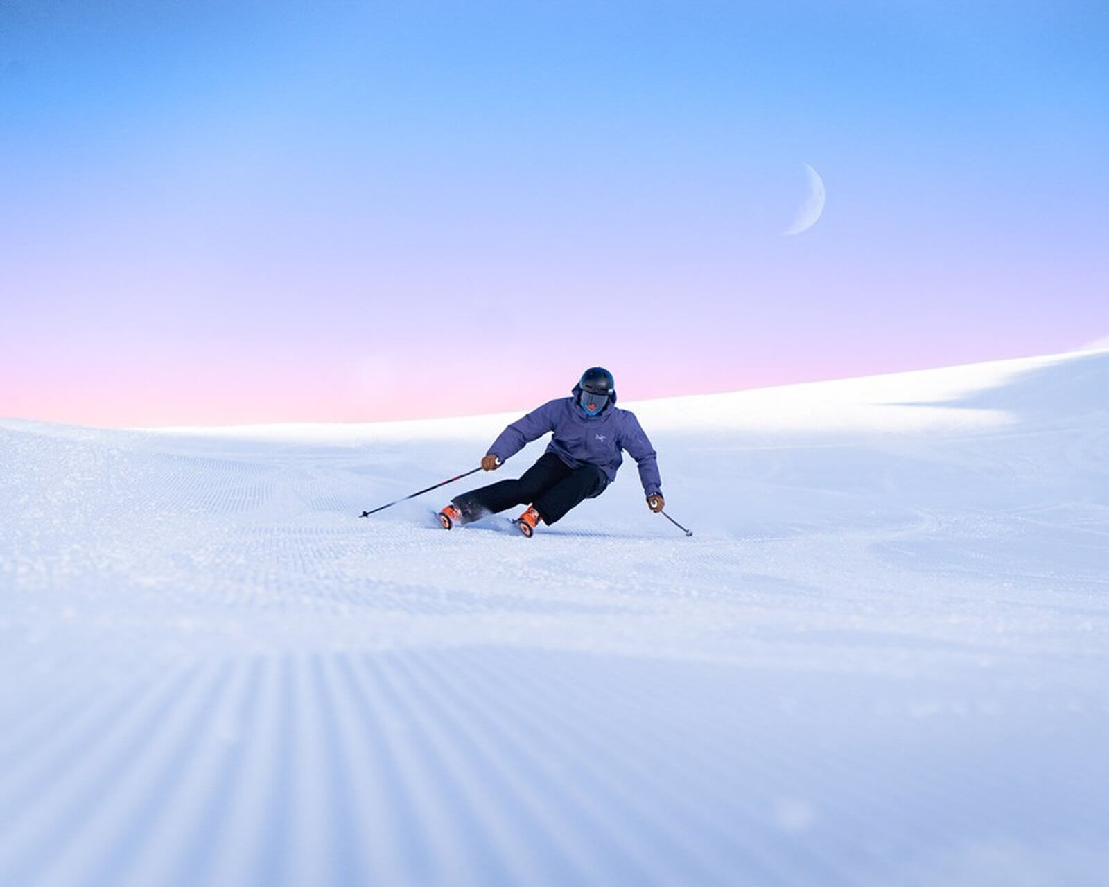 Skier carving turns on fresh snow under a colorful sunrise sky with a crescent moon visible.
