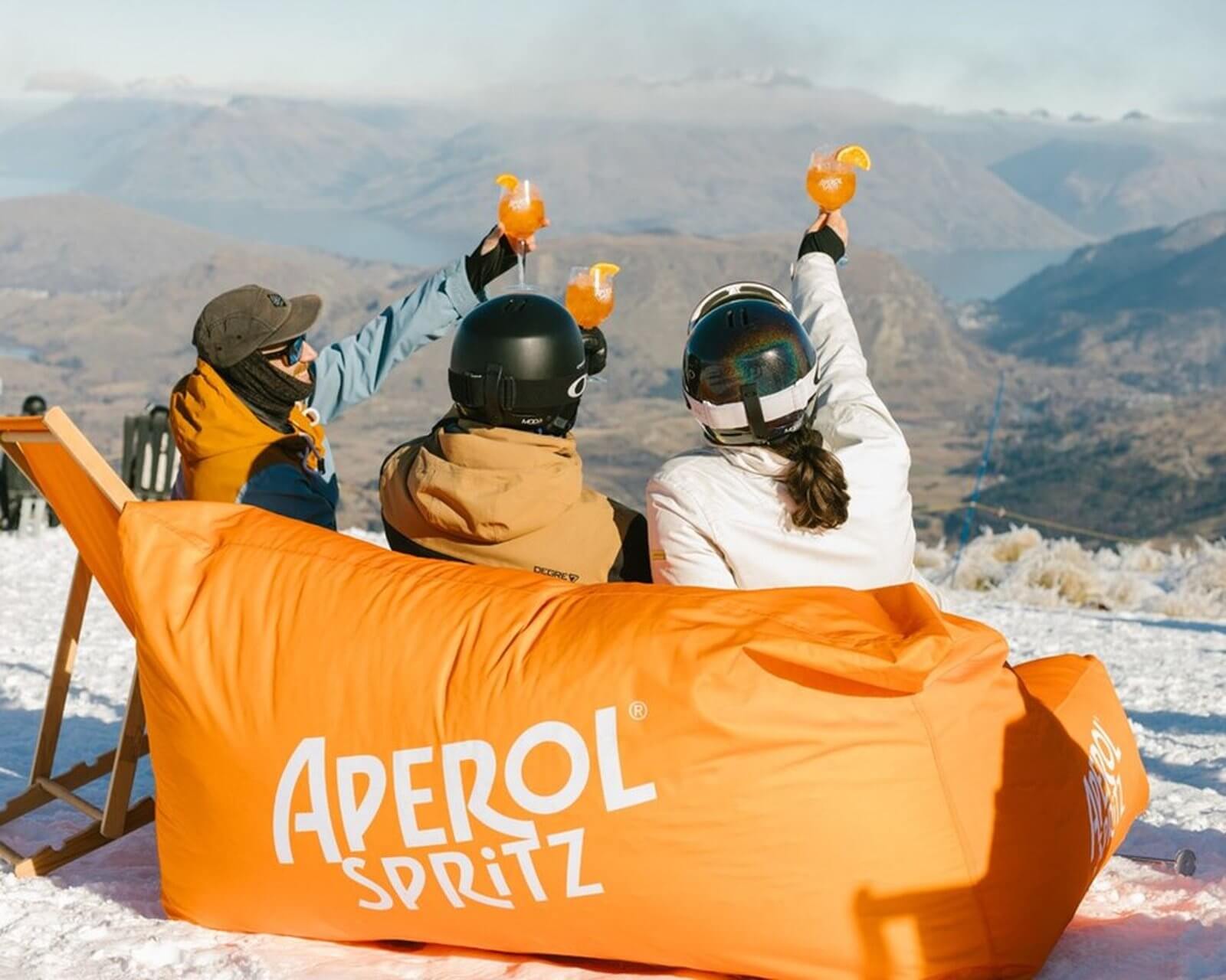 Three people in winter gear raising Aperol Spritz glasses while sitting on an orange branded couch in snow.