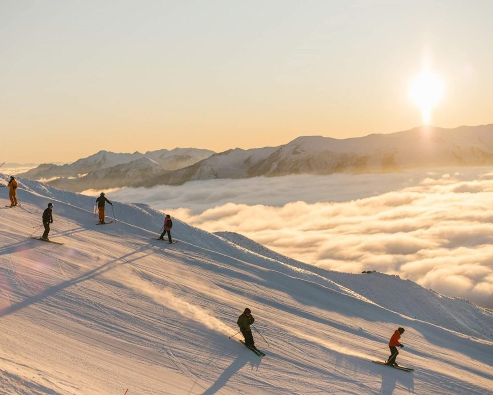 Skiers descending a snowy mountain slope at sunset above clouds with distant mountains in view.