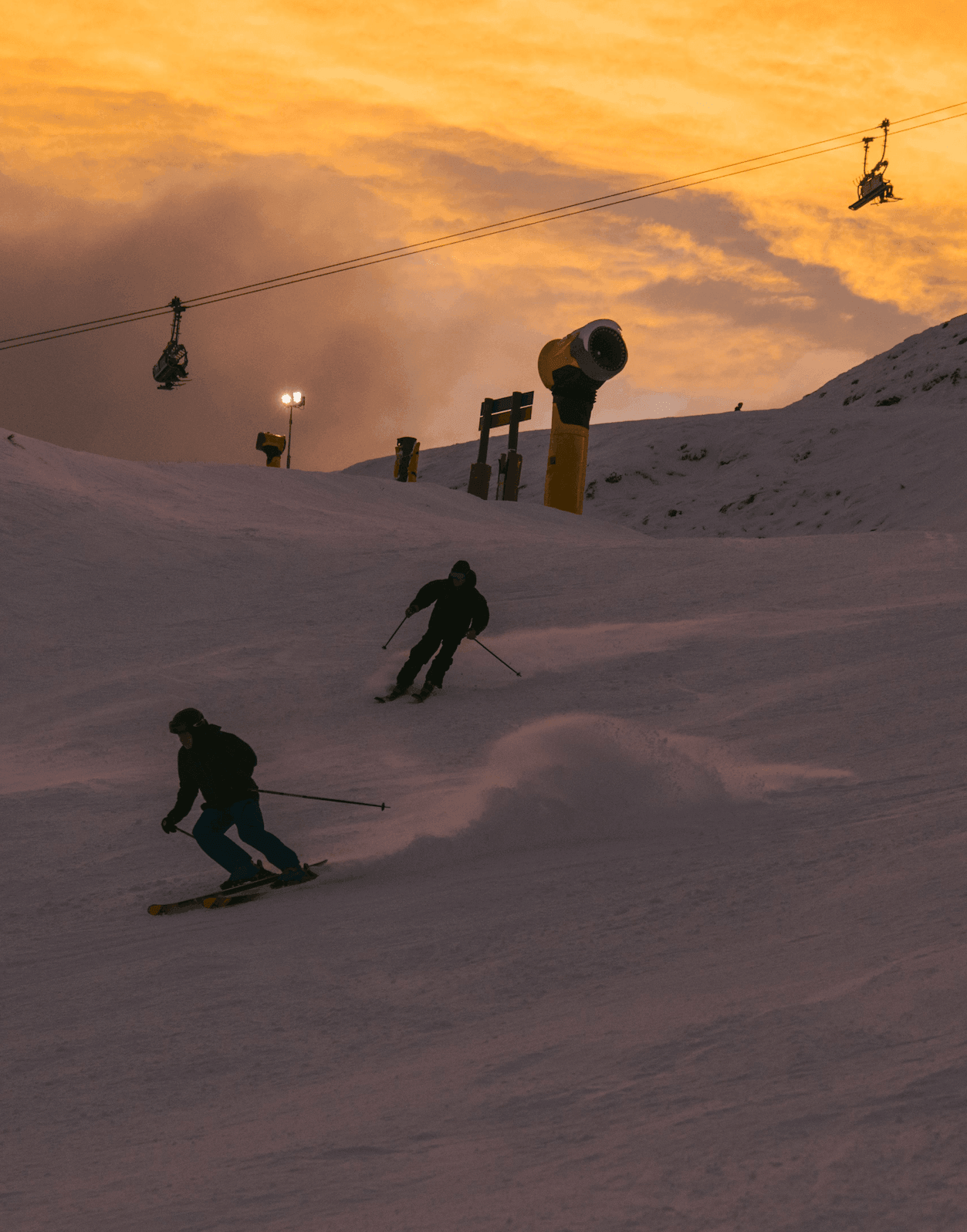 Two skiers descend a snowy slope at sunset beneath chairlifts and snow machines.
