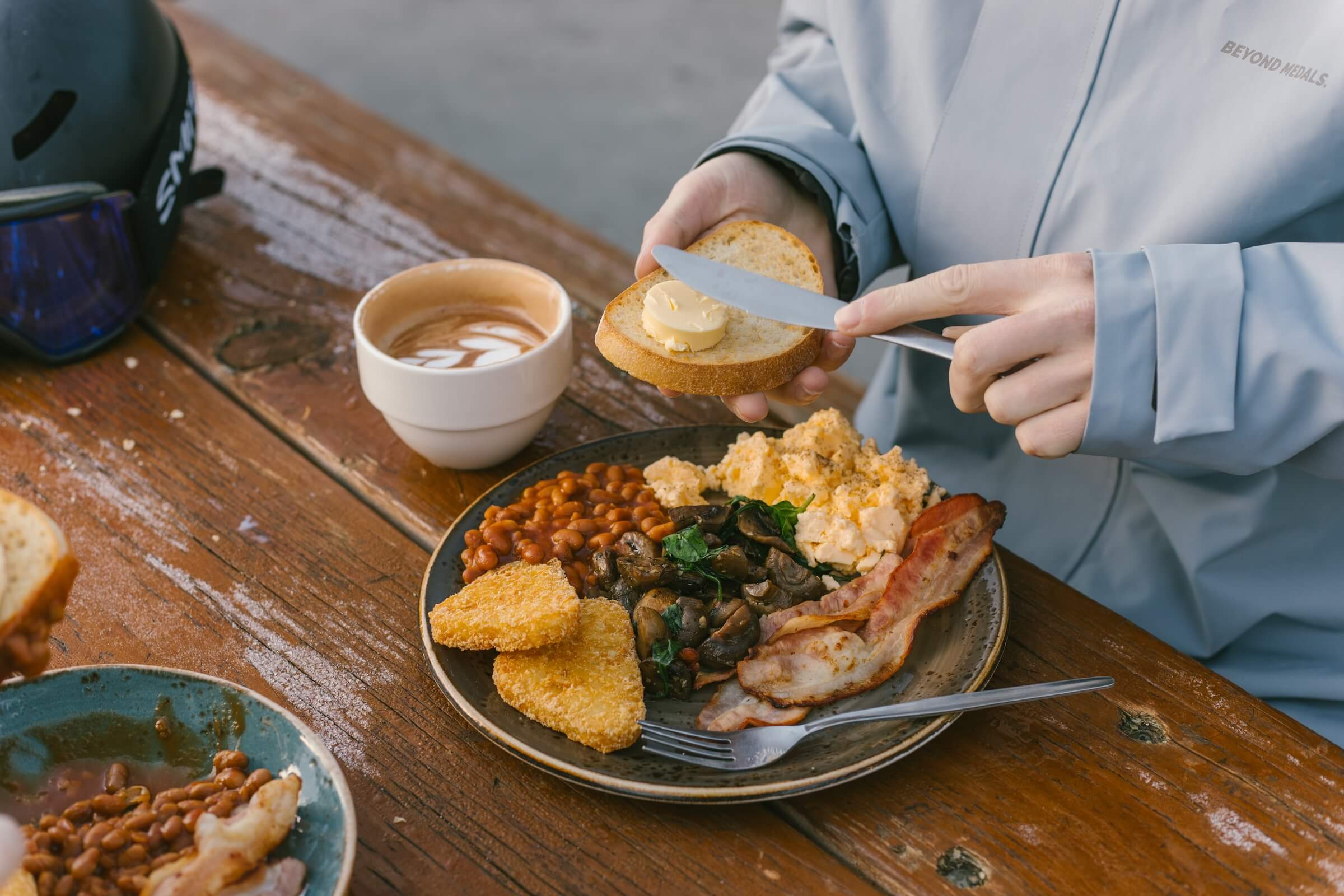 Person spreading butter on toast beside a plate with eggs, bacon, mushrooms, beans, and hash browns.