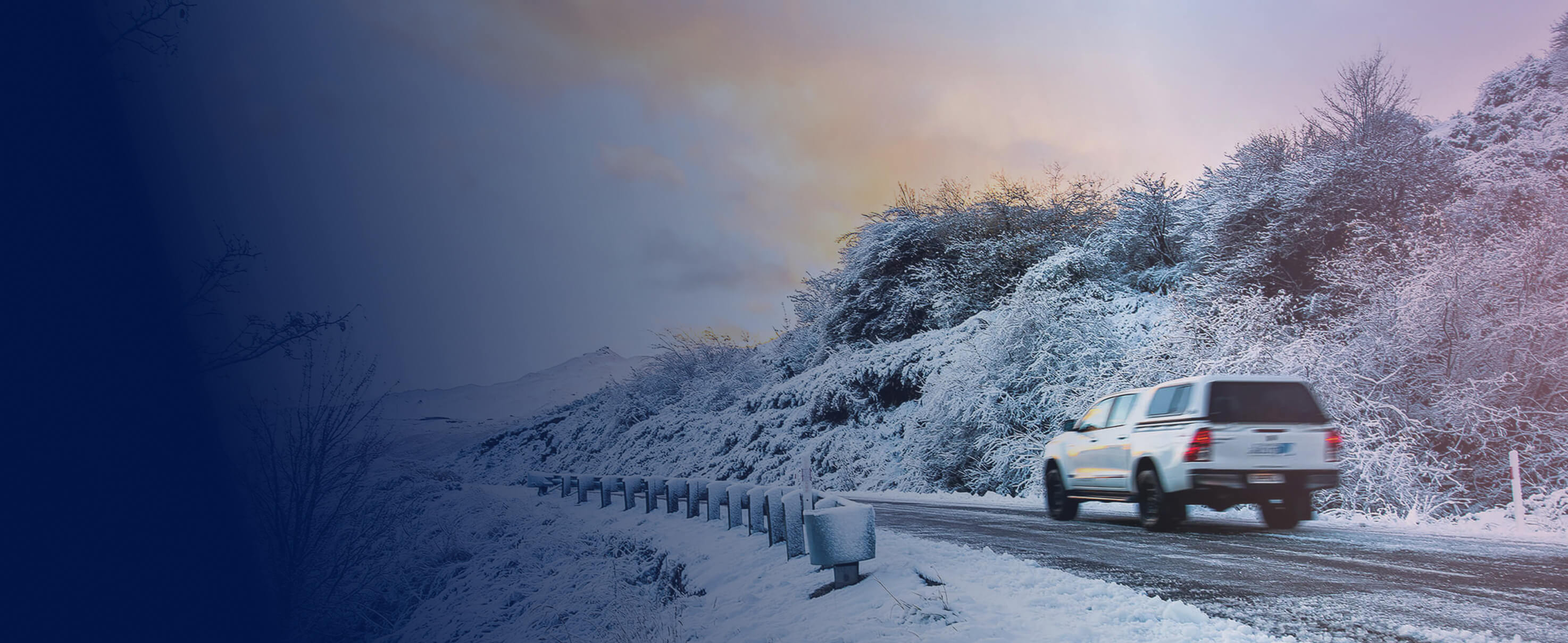 White pickup truck driving on a snowy mountain road surrounded by snow-covered trees at sunset.