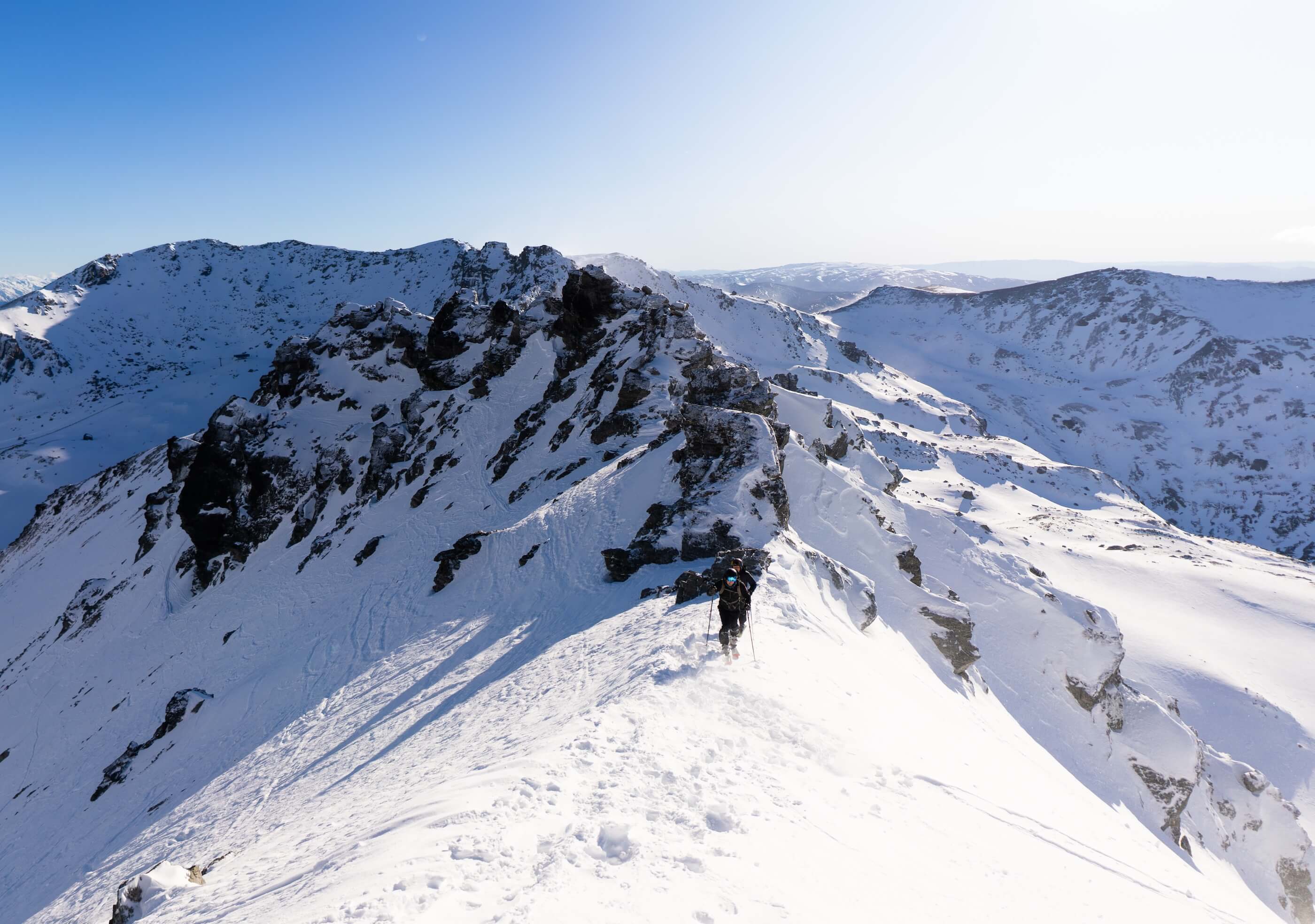 Hikers wearing snow gear and masks trekking on snowy mountain ridge under clear blue sky.