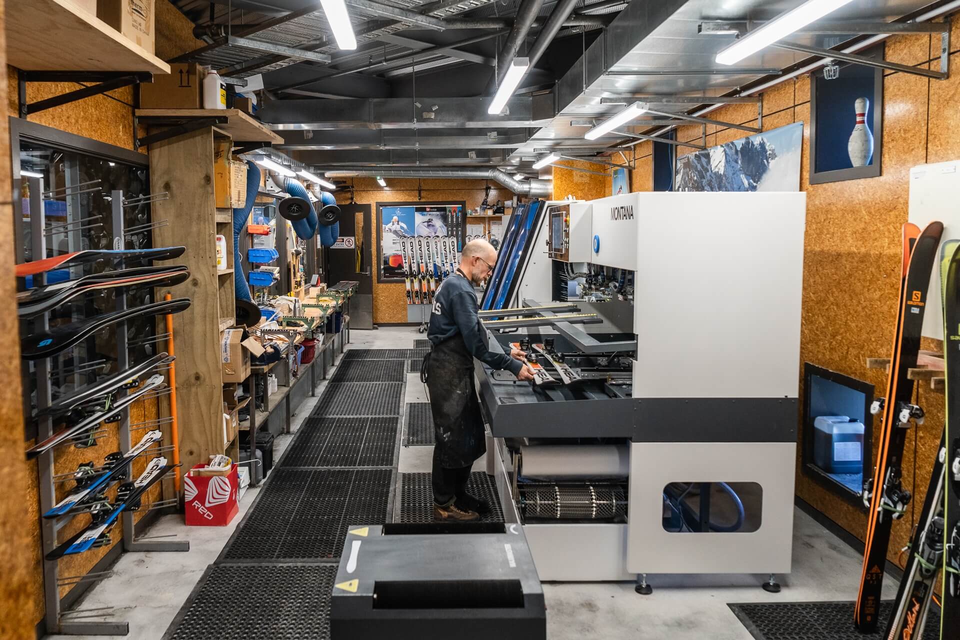 Person working on a Montana ski machine in a workshop filled with skis and ski equipment.