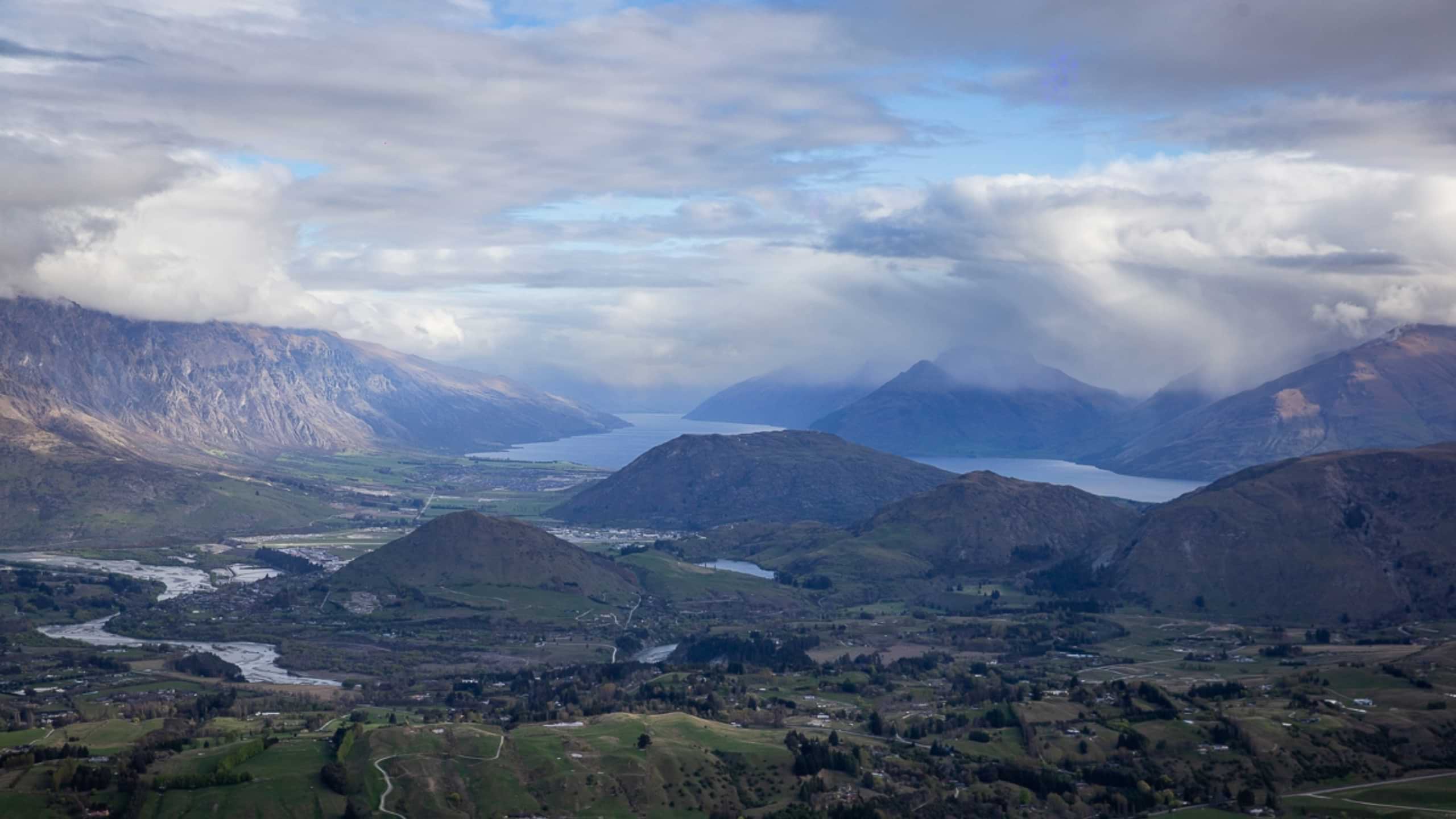 Valley with green fields, winding rivers, lakes, and mountains under a cloudy sky.