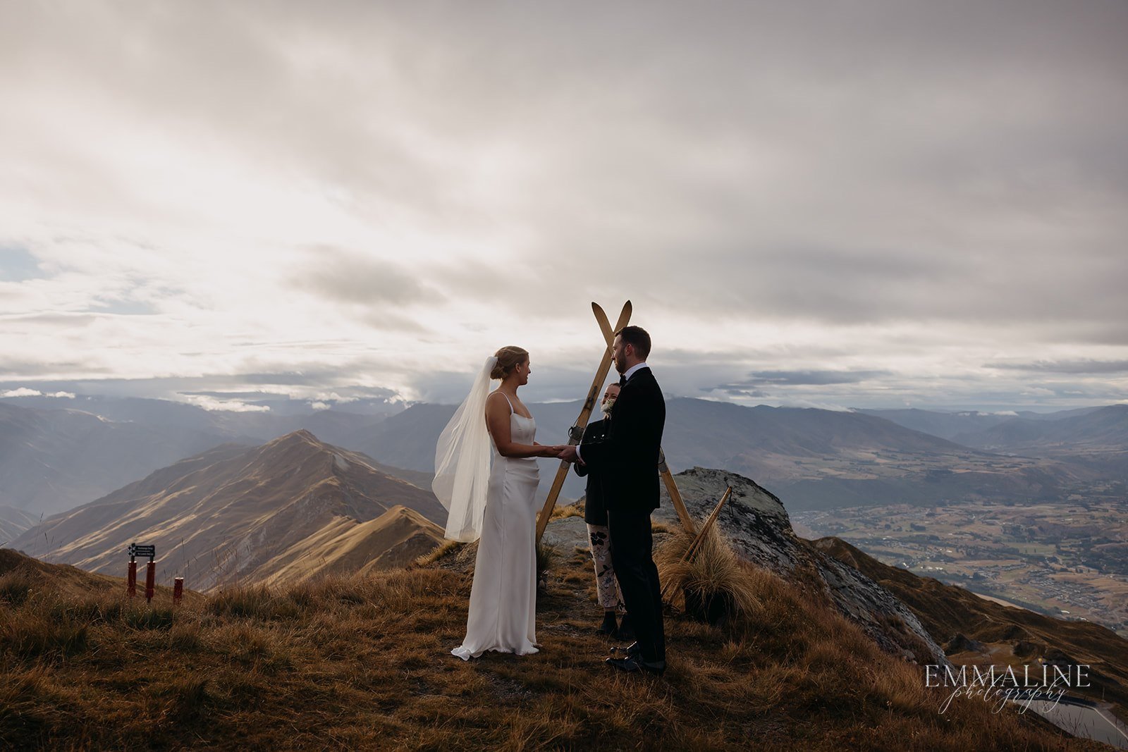Bride and groom holding hands on mountain top during outdoor wedding ceremony at sunset.
