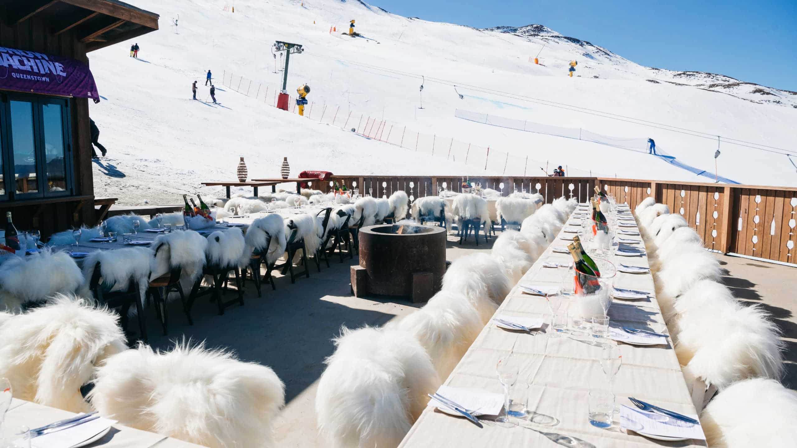 Outdoor dining tables with white fur-covered chairs set against a snowy ski slope backdrop.