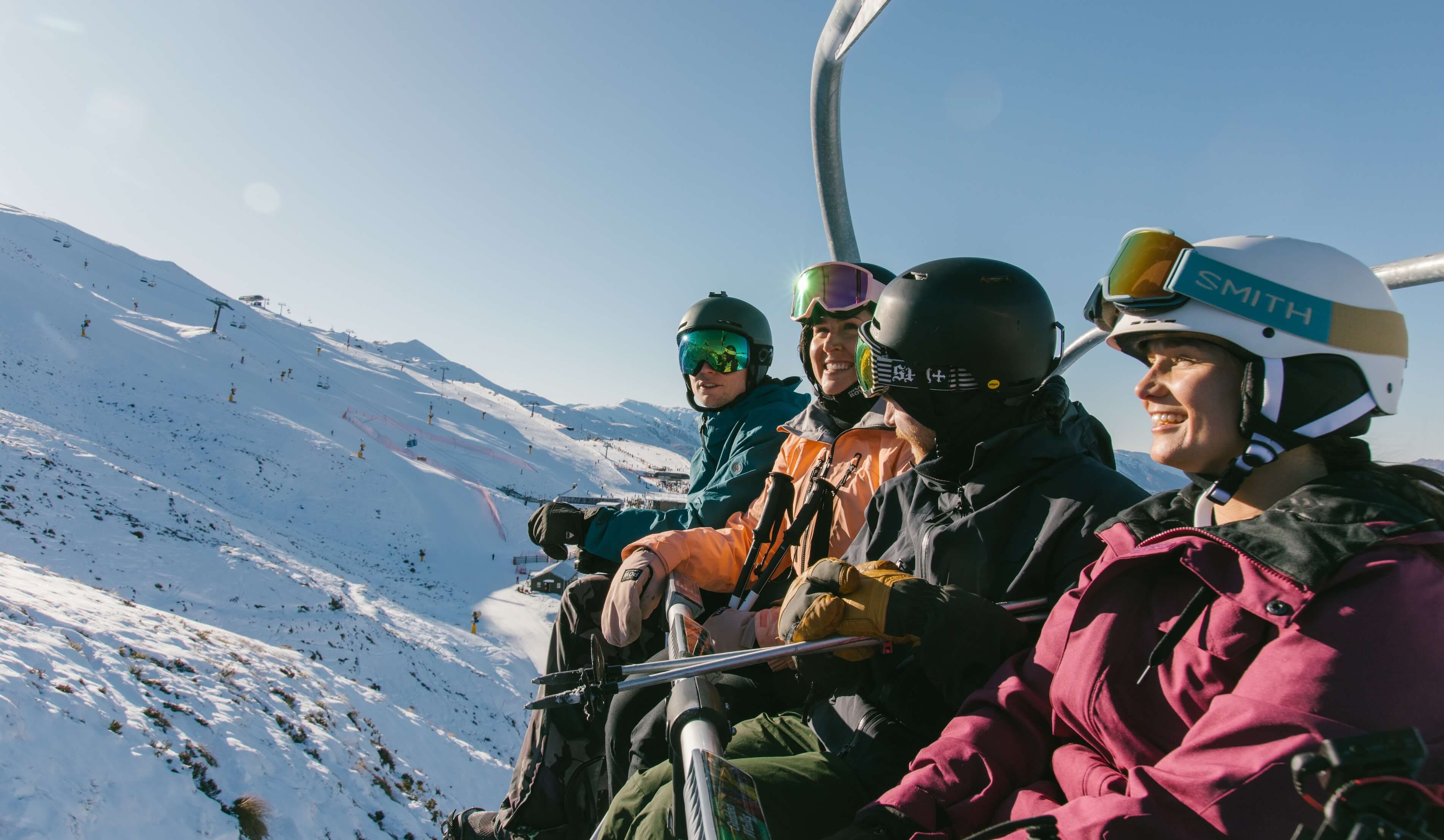 Four skiers in helmets and goggles riding a ski lift over snowy slopes on a sunny day.