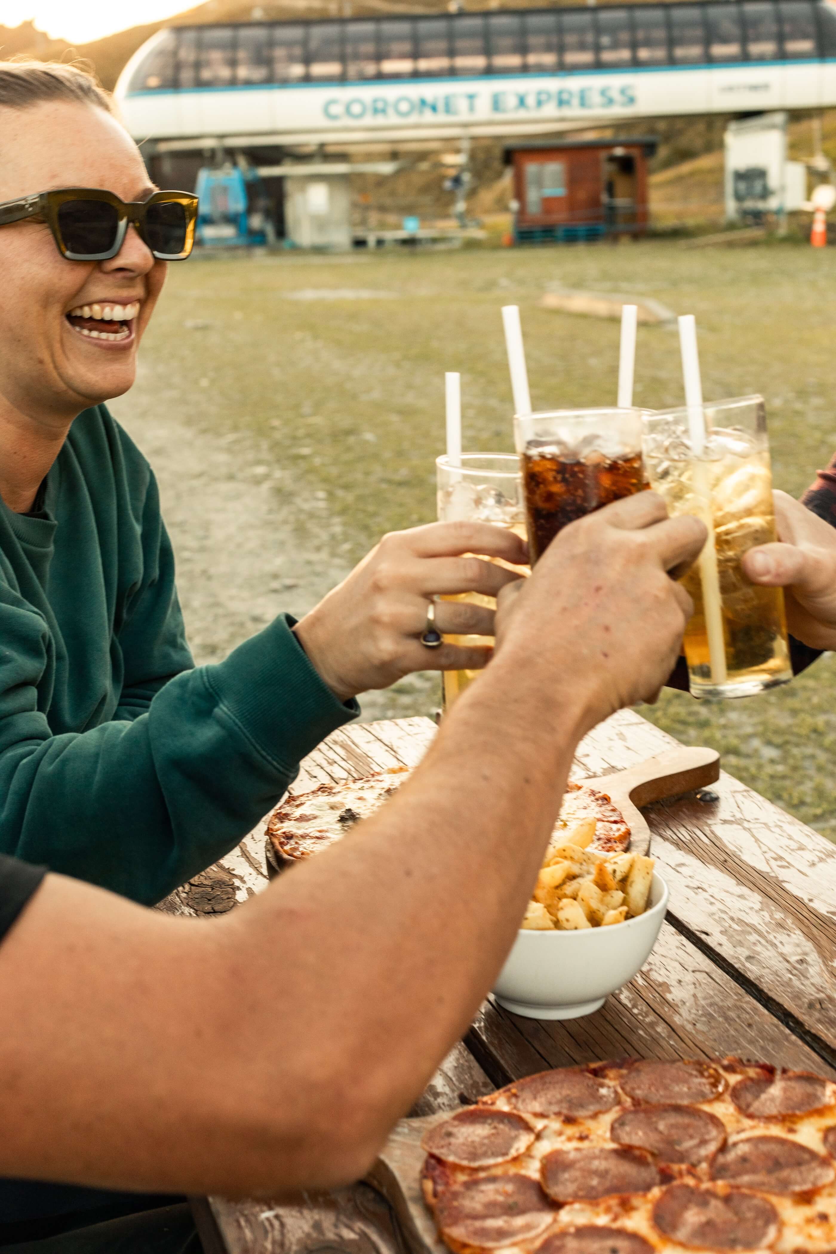 Friends clinking drinks outdoors at a picnic table with pepperoni pizza and fries.