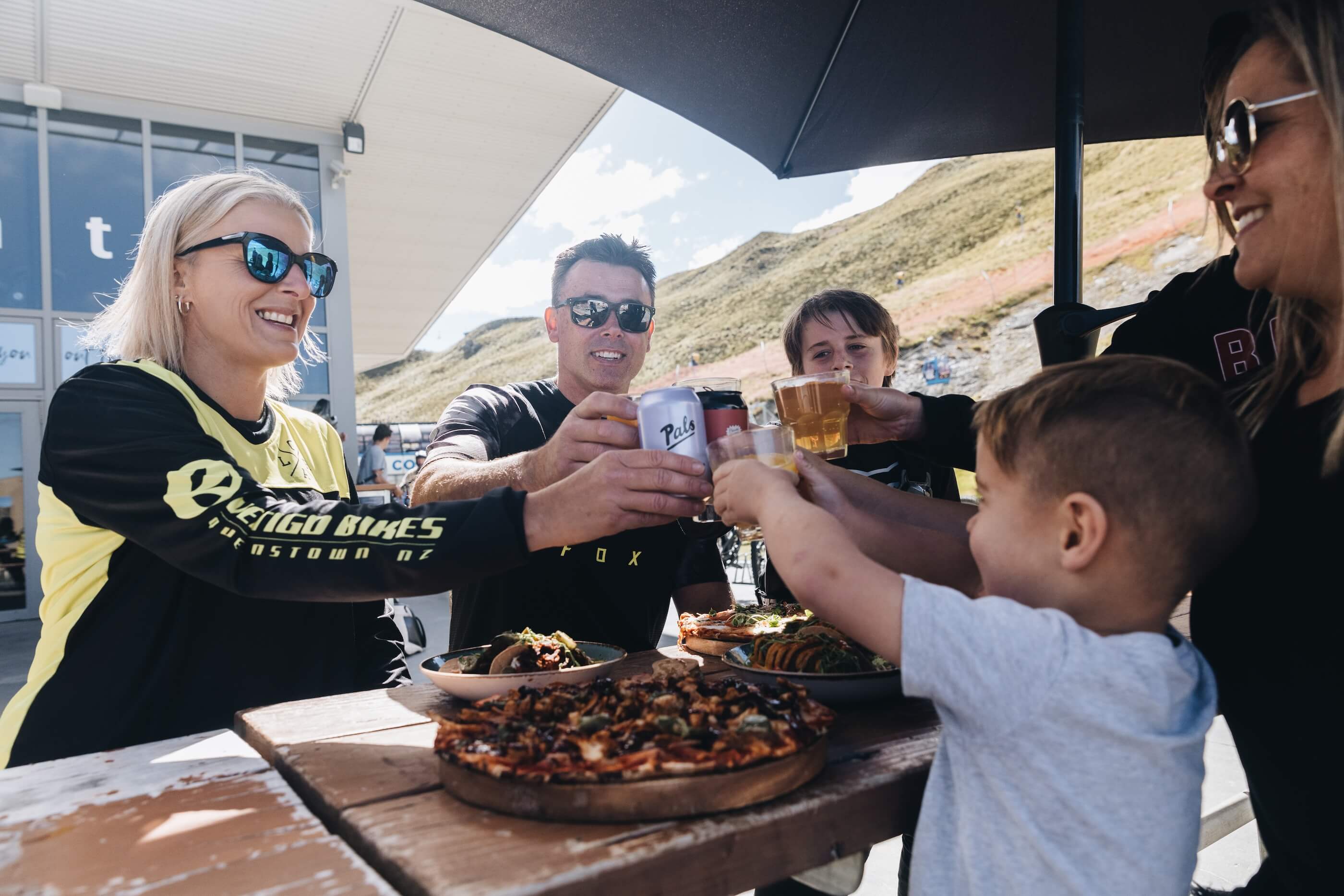 Group of people outdoors clinking drinks over a table with pizza and food under umbrella.