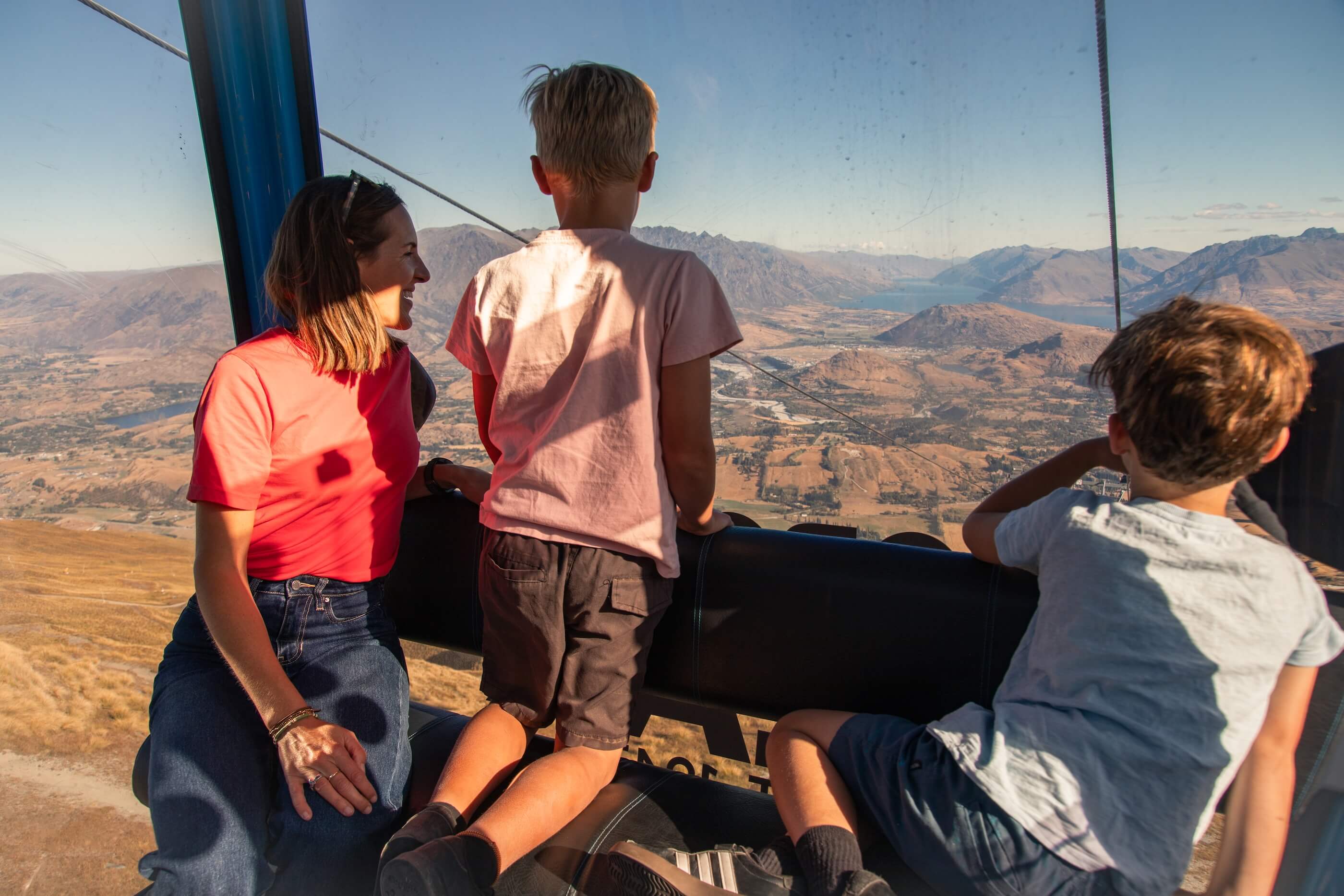 Woman and two boys enjoying mountain views from inside a cable car on a sunny day.