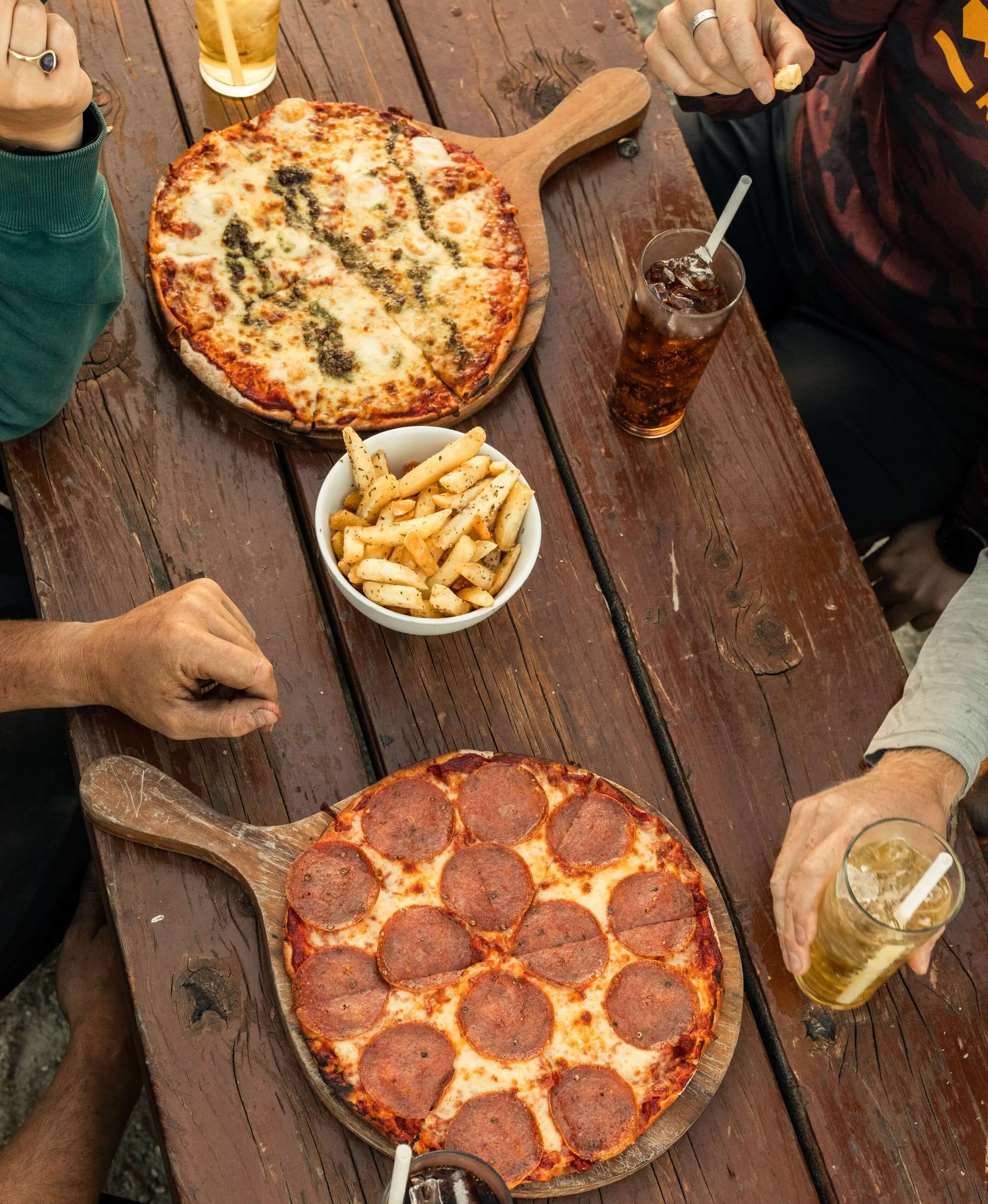 Two pizzas, a bowl of seasoned fries, and four drinks on a wooden picnic table with people’s hands.