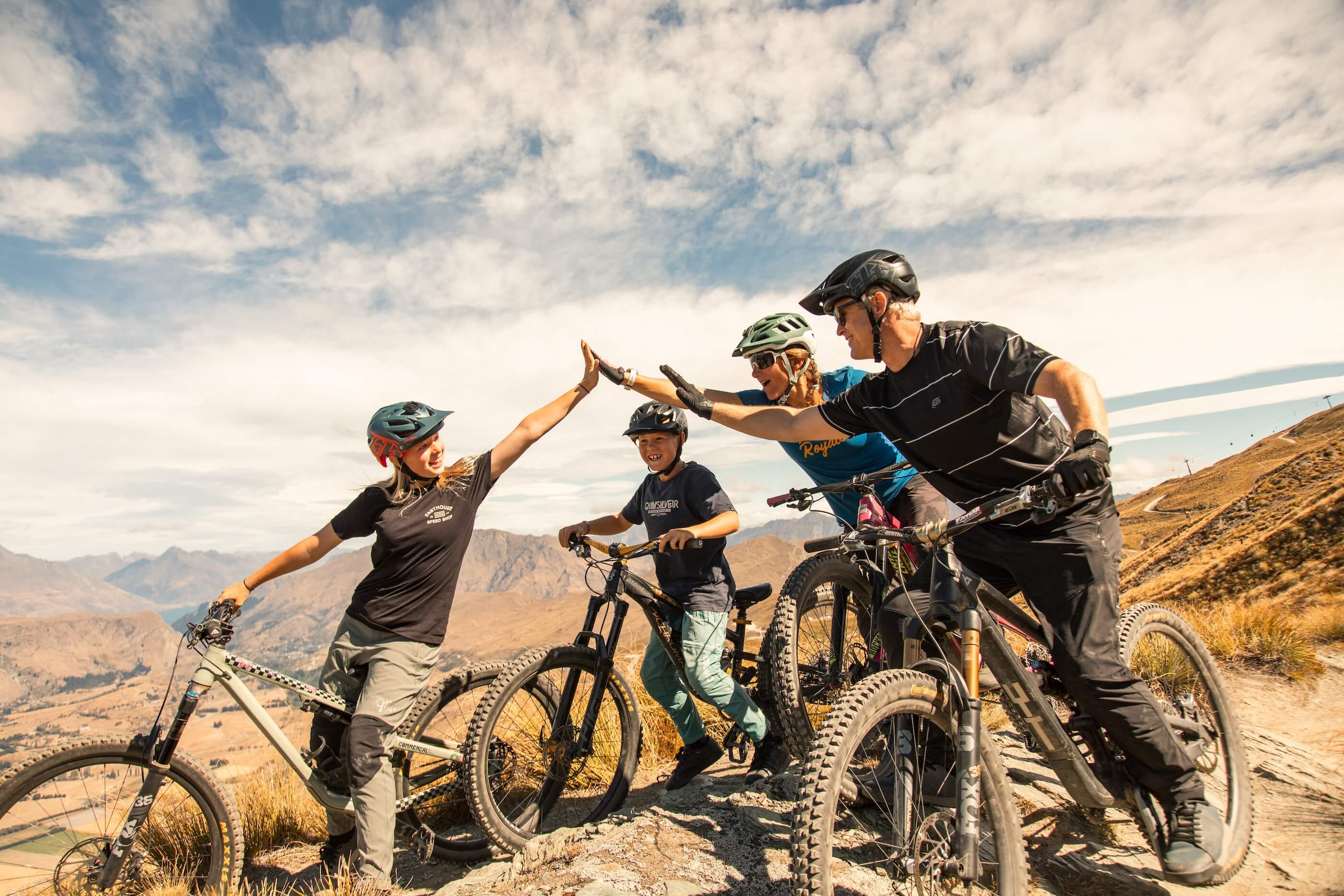 Four mountain bikers on a rocky trail giving high fives with mountains and sky in the background.