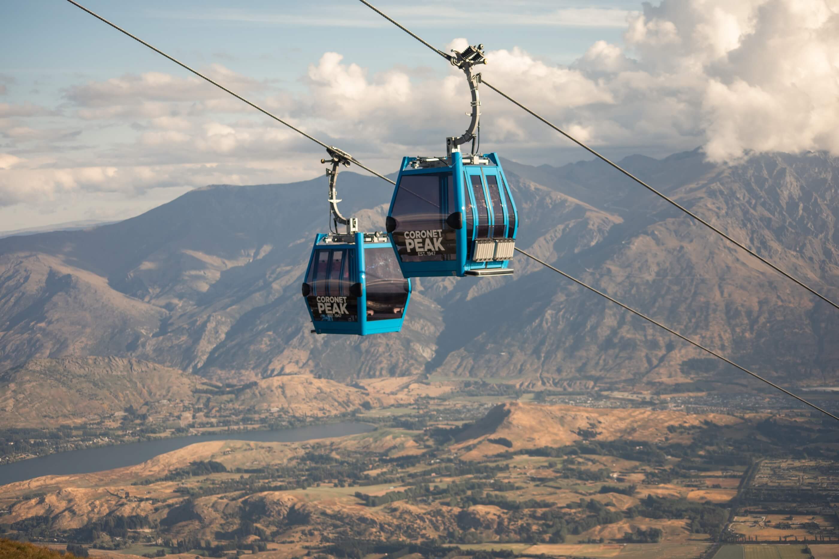 Two blue Coronet Peak gondolas suspended on cables above a mountainous landscape with a lake below.
