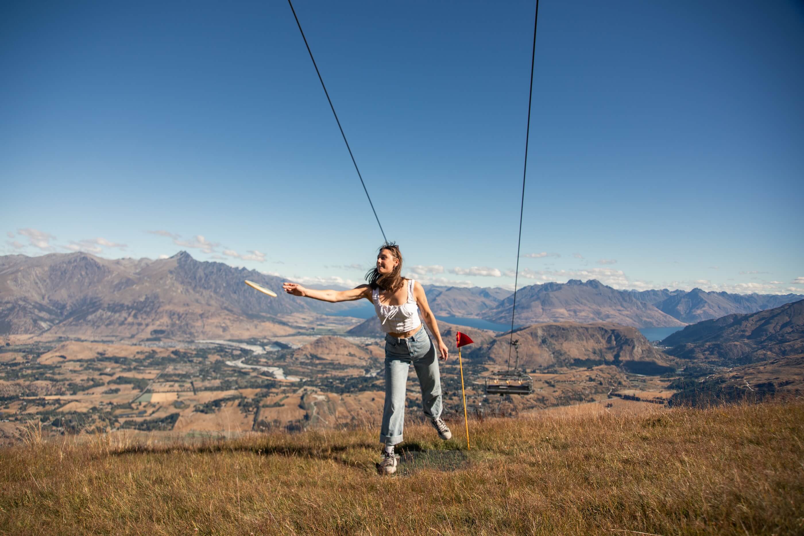 Woman throwing a frisbee on a grassy hill with mountains and a clear blue sky in the background.