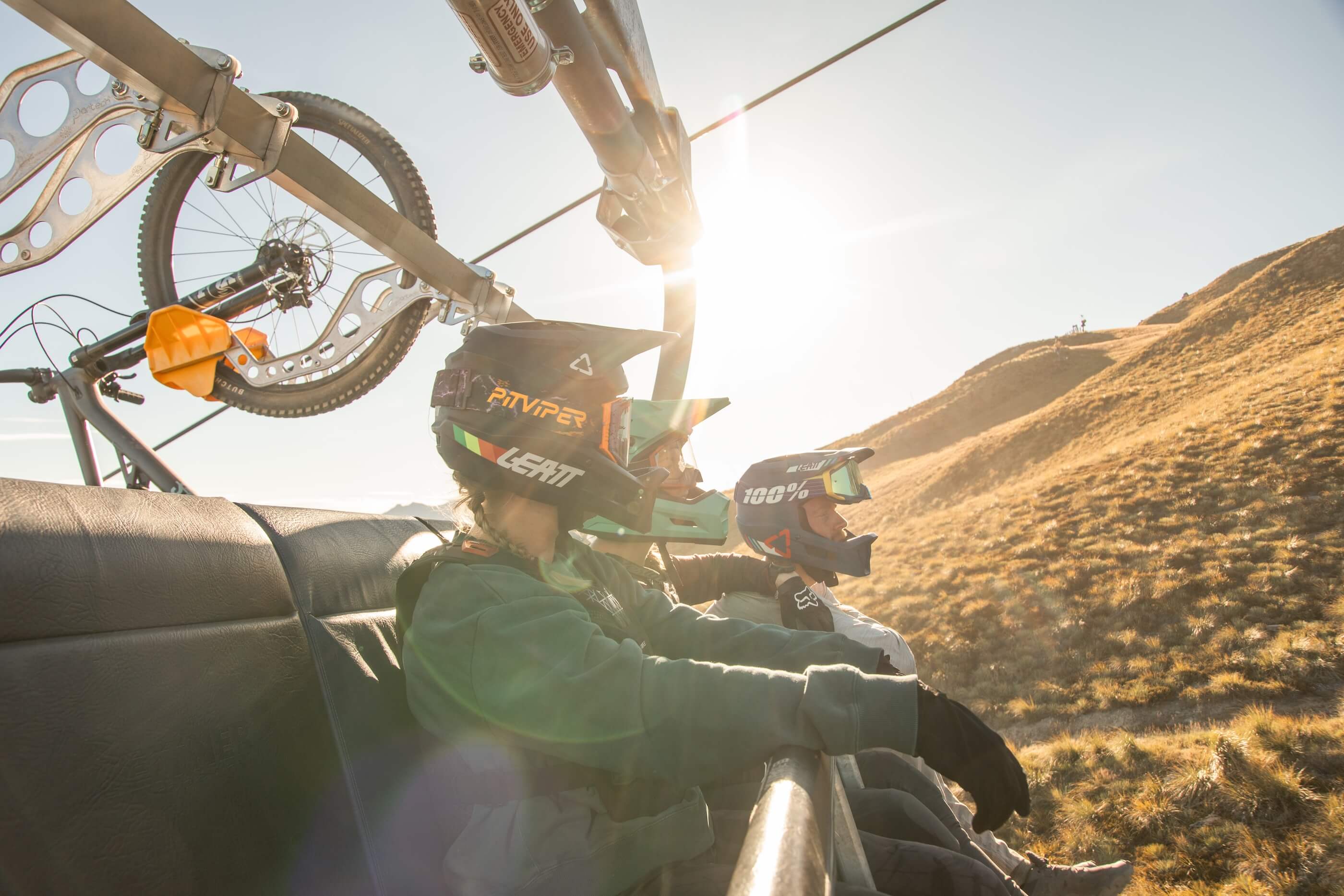 Three mountain bikers wearing helmets sit on a chairlift with a bike hanging above, riding uphill.