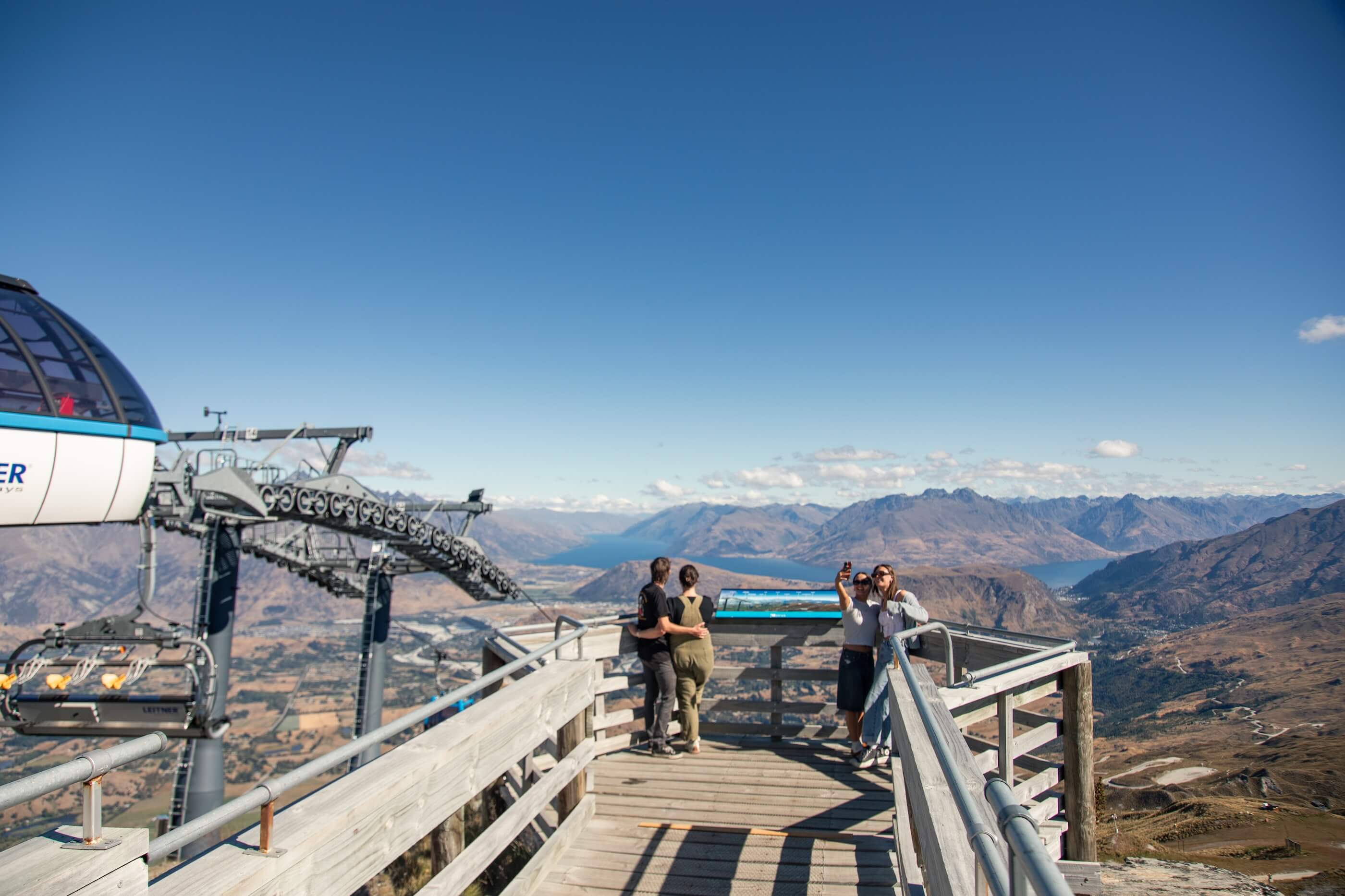 Four people on a wooden lookout with mountain and lake views, two taking a selfie, others admiring scenery.
