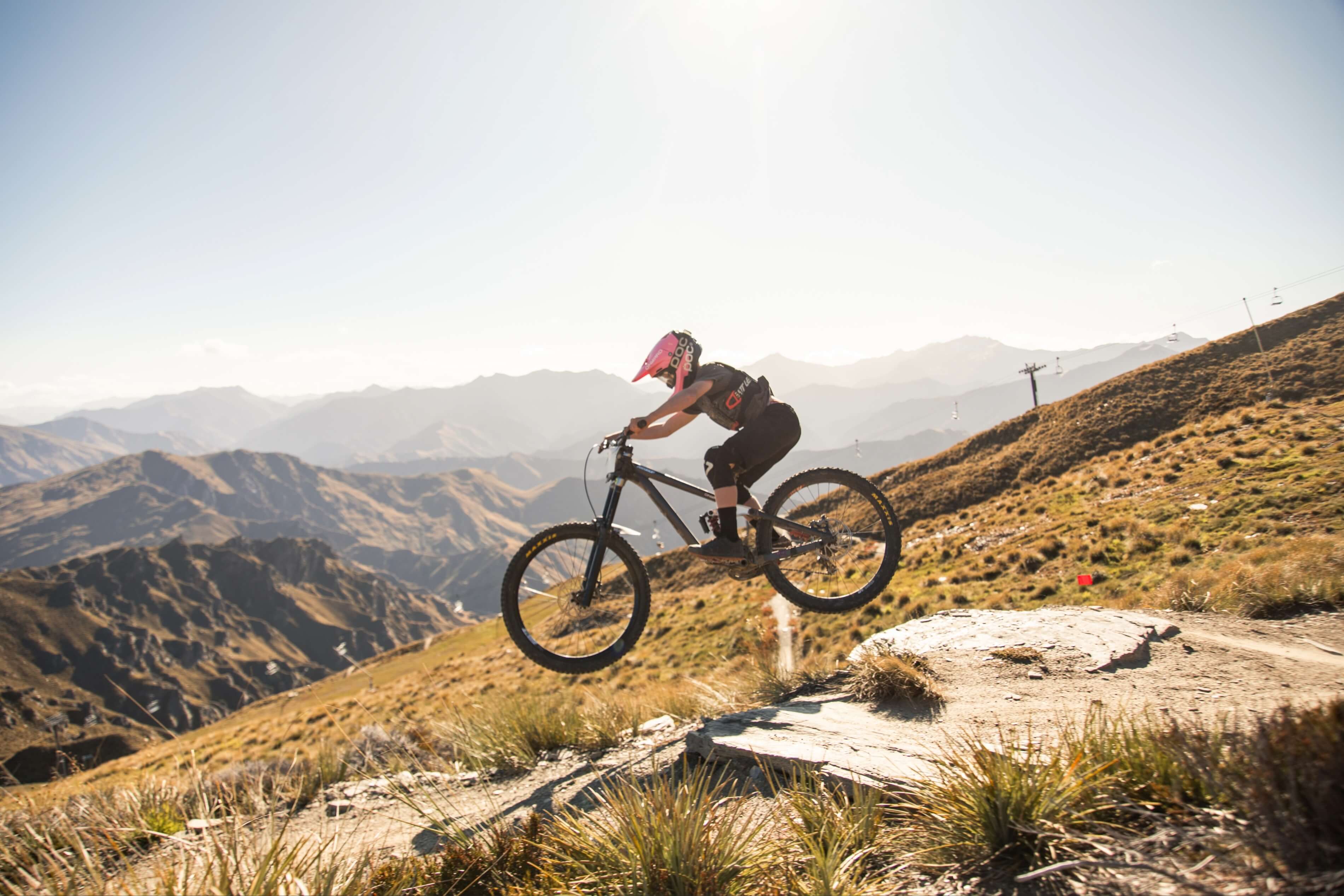 Mountain biker wearing a pink helmet jumps over a rocky trail with mountains in the background.