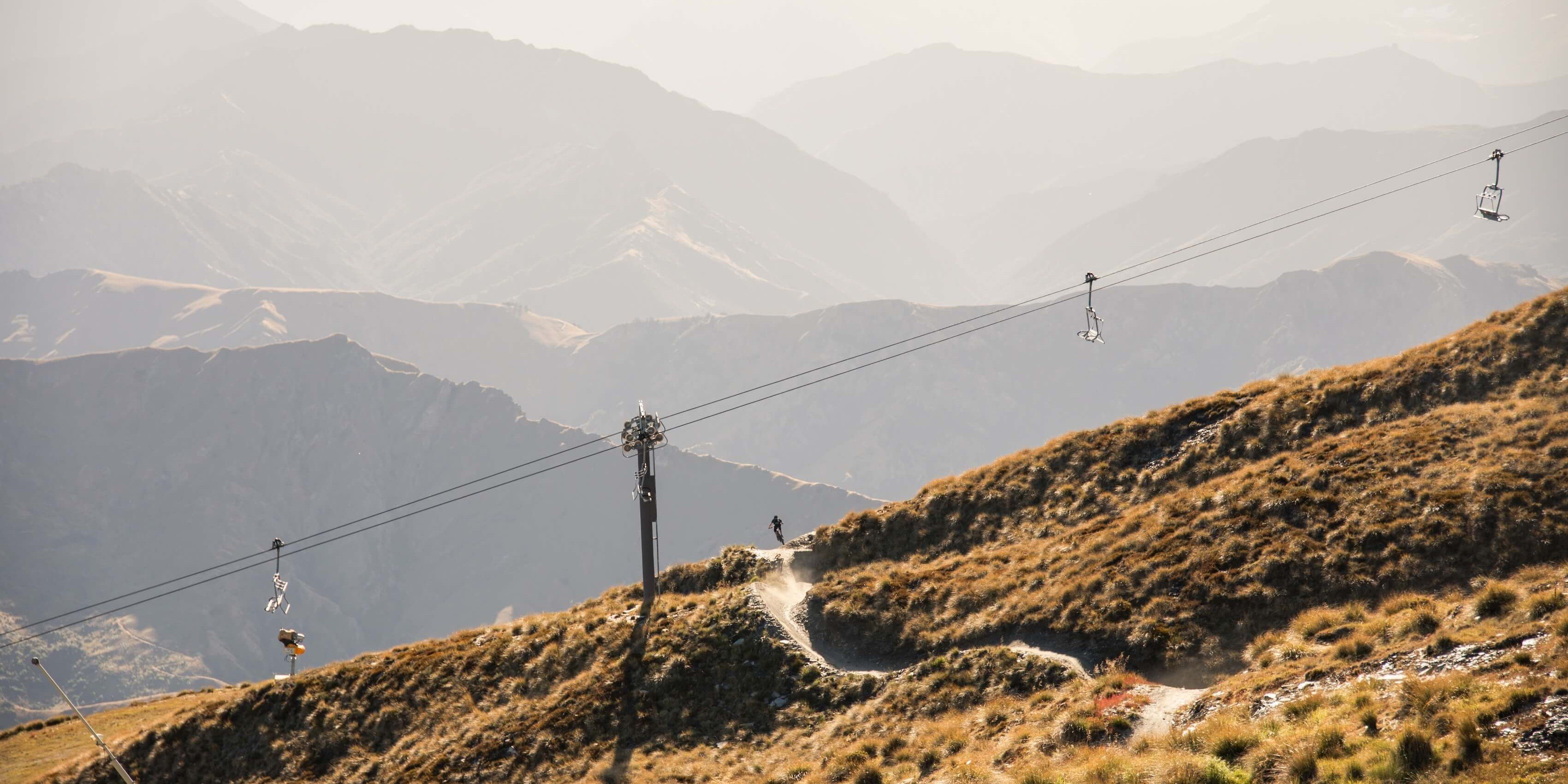 Mountain biker riding on a dusty trail beneath ski lift cables and chairs in a mountainous landscape.
