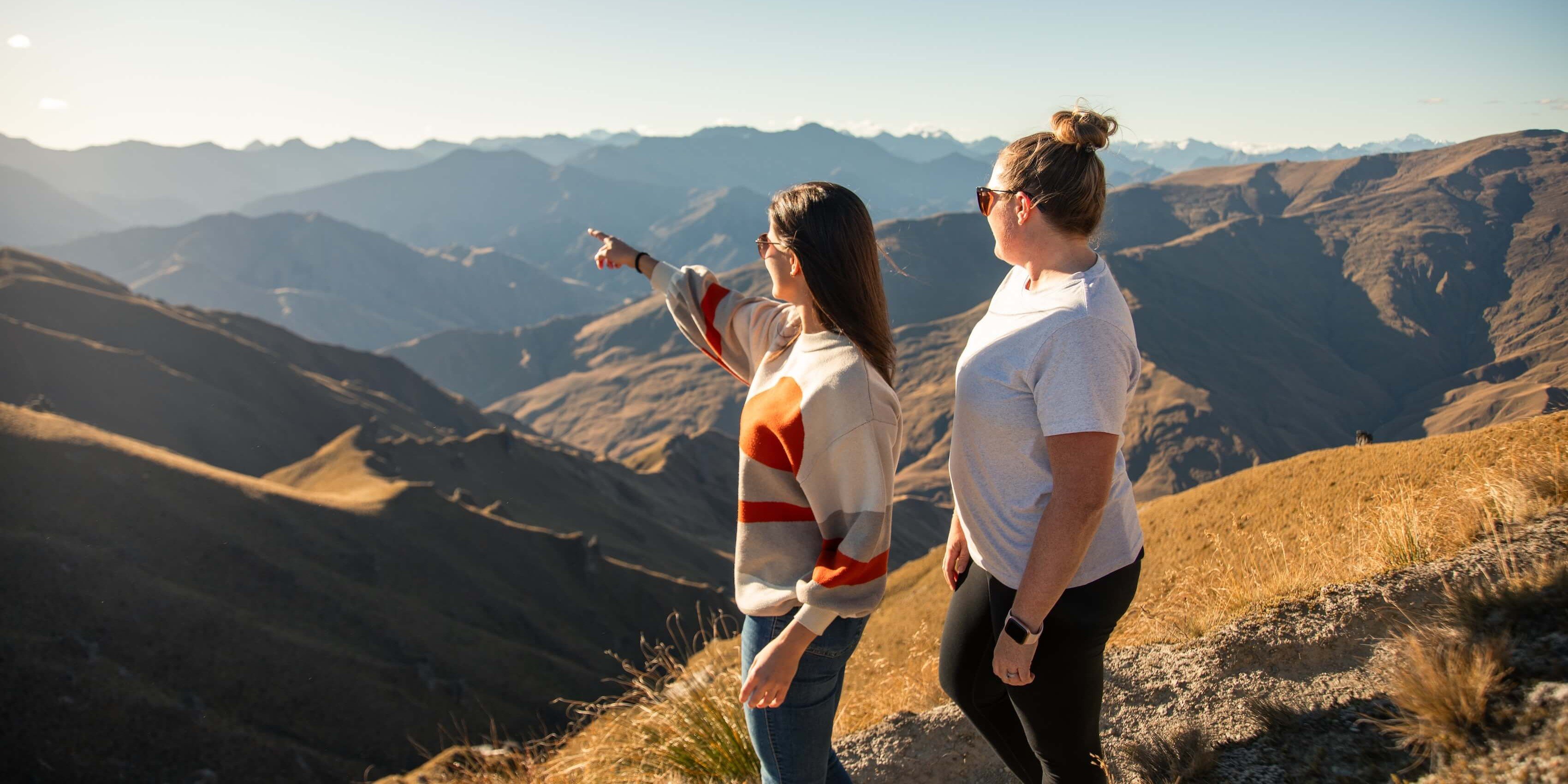 Two women wearing sunglasses stand on a mountain path looking at distant mountain ranges.