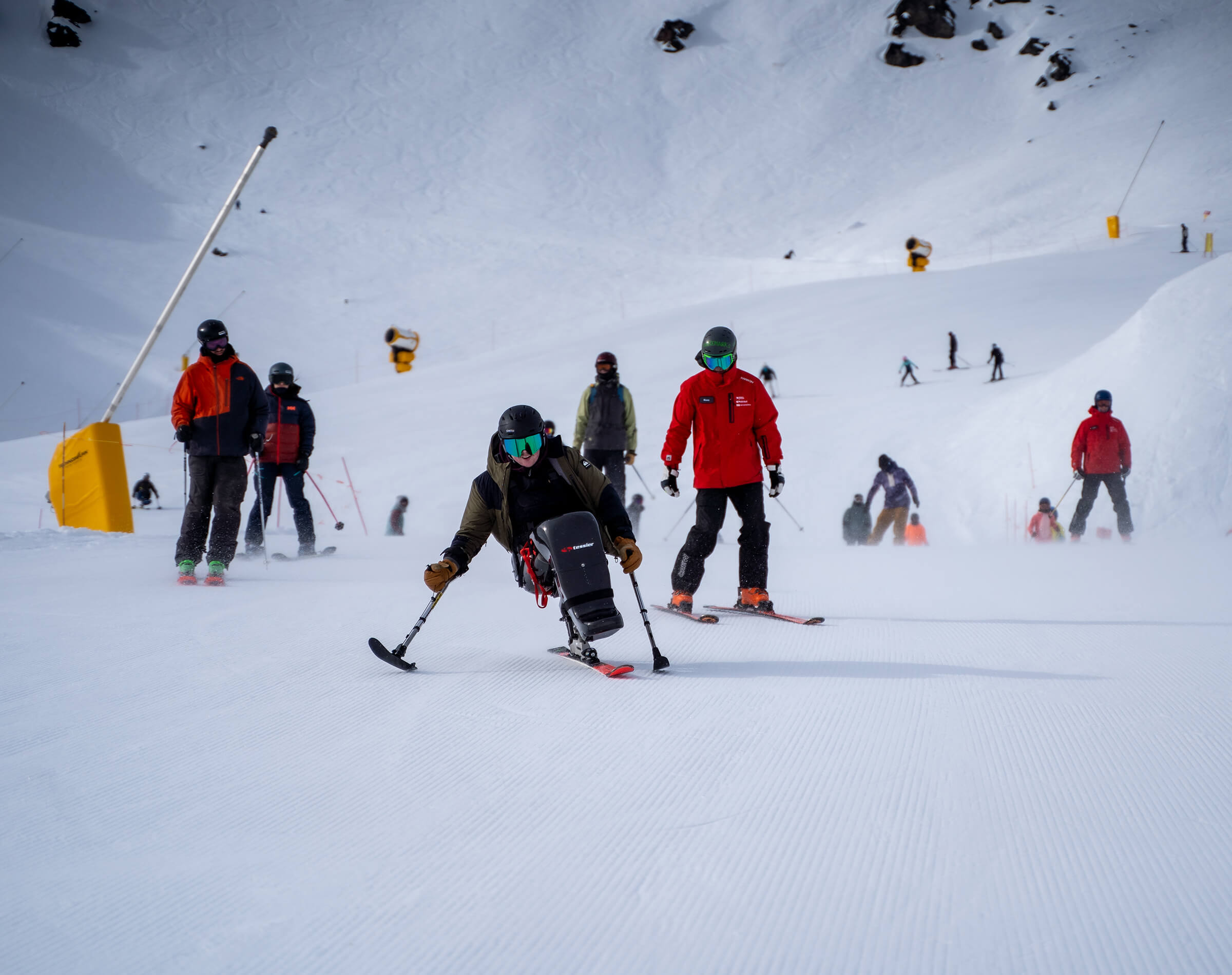 Person skiing on adaptive sitski with others skiing on snowy mountain slope.