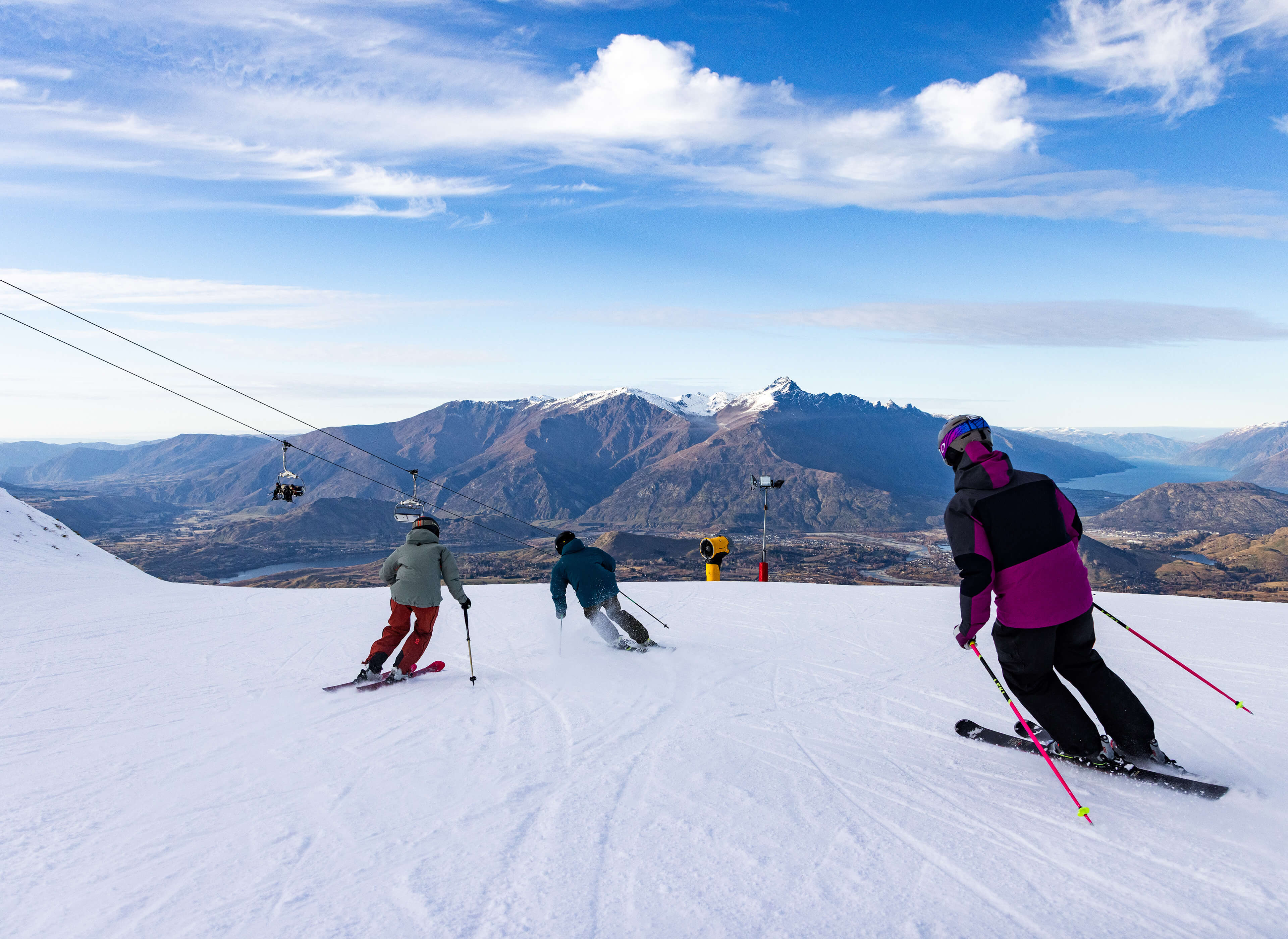 Three skiers descending a snowy slope with mountain and lake views on a clear day.