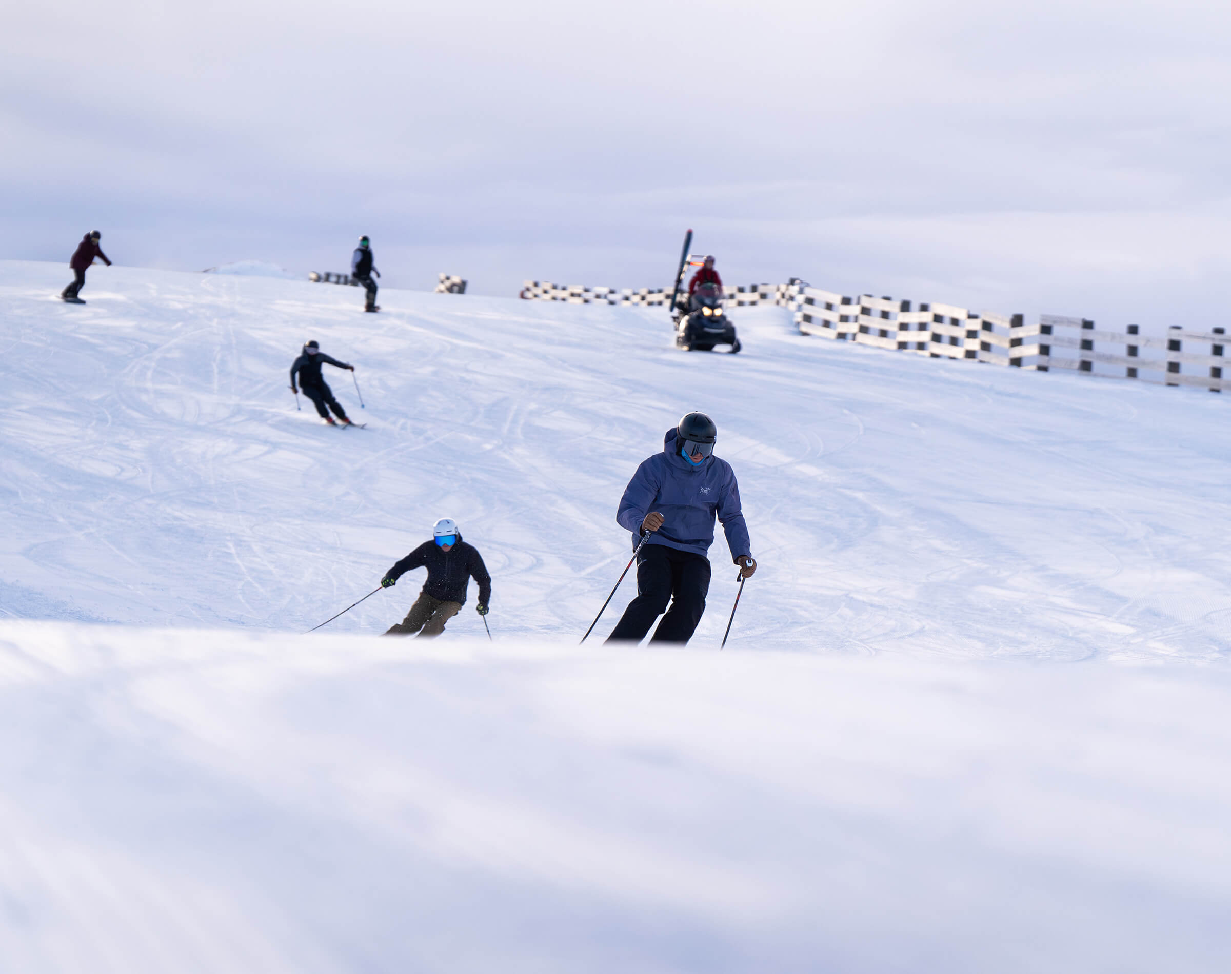 Multiple skiers and snowboarders on a snowy slope with a snowmobile in the background.