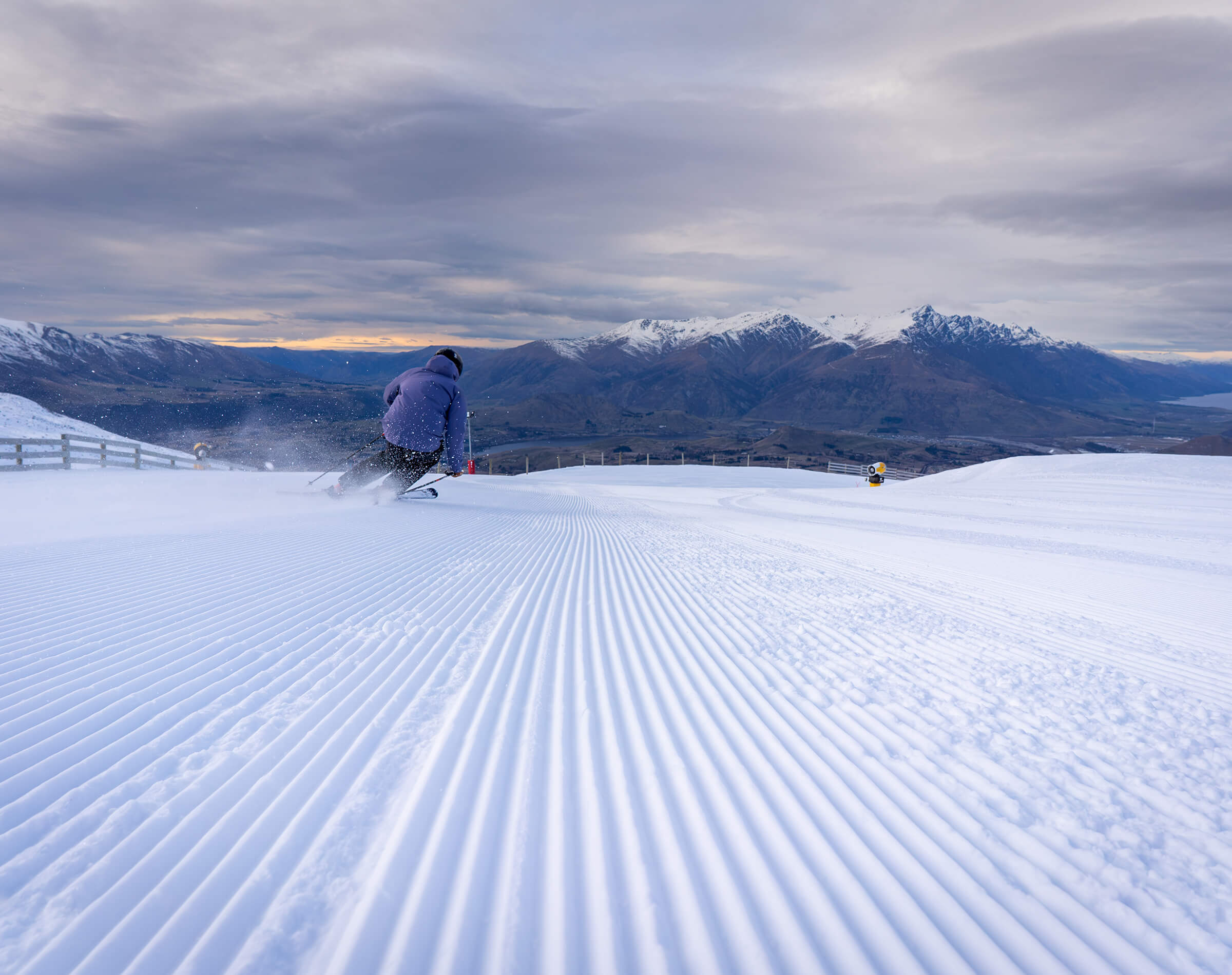 Skier carving turns on freshly groomed snow with snowy mountains and cloudy sky in the background.