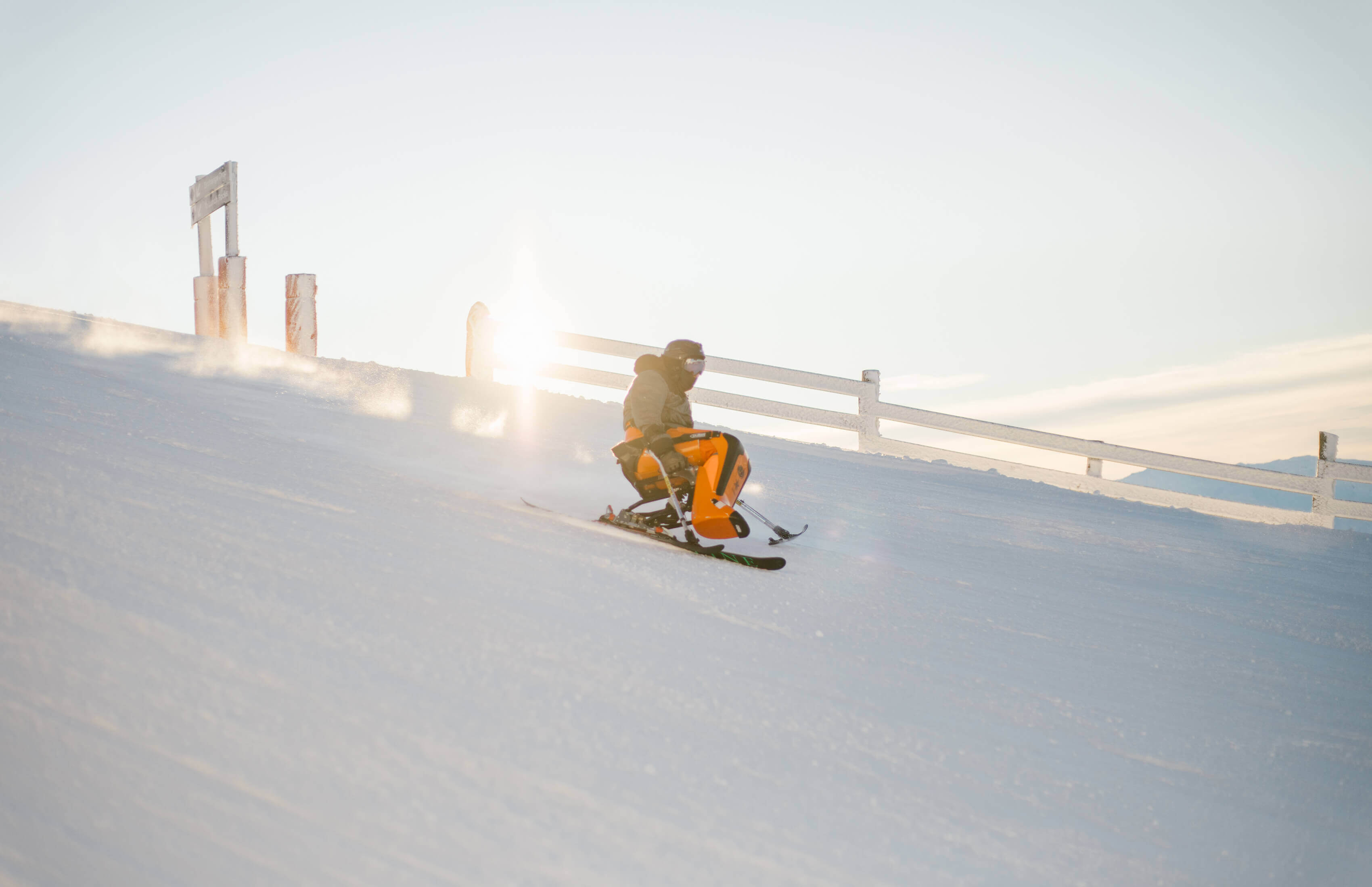 Person skiing downhill in a sit-ski on snow with sunlight behind a white fence.