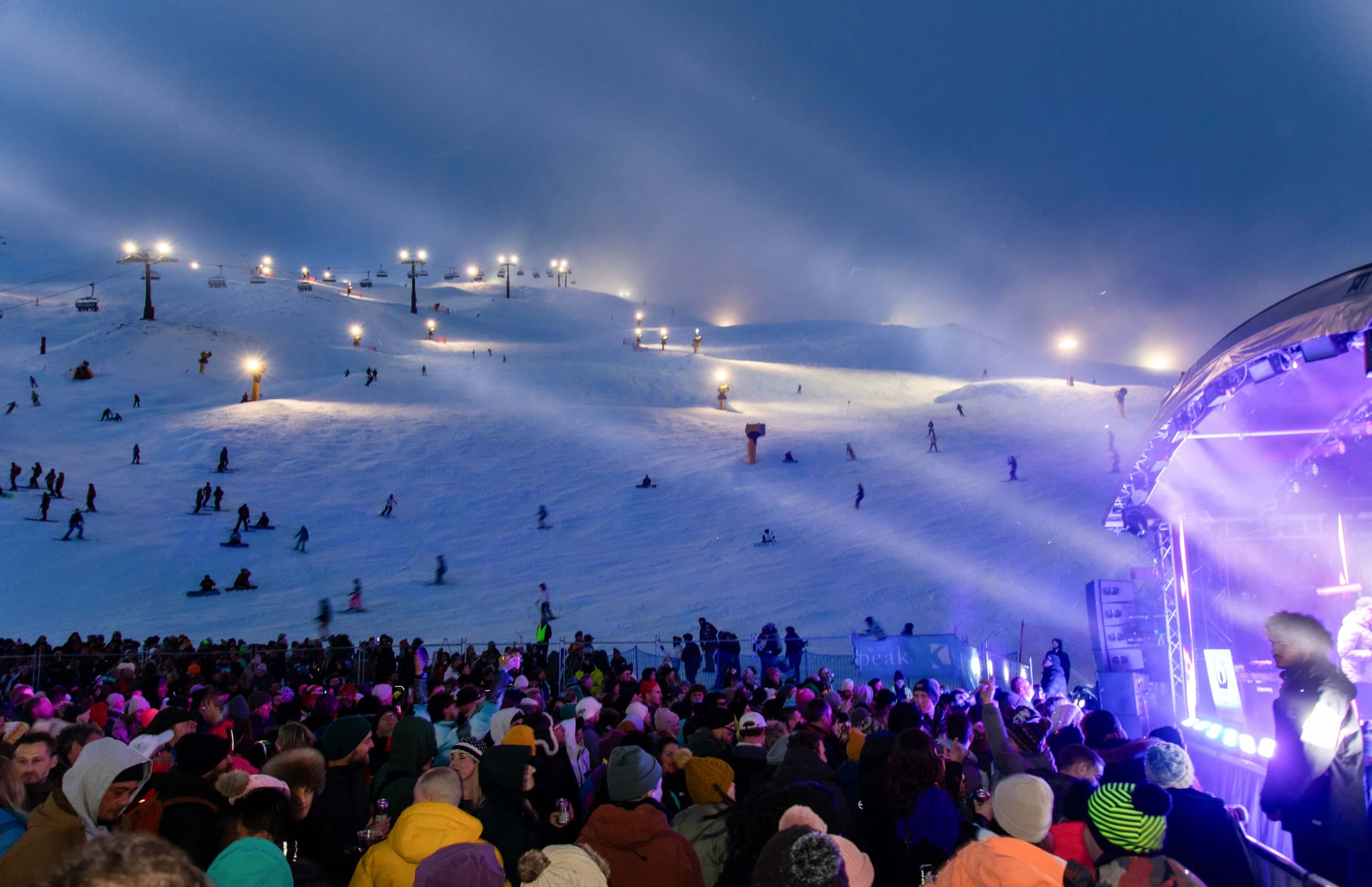 Crowd at night ski party near snowy slopes lit by bright lights and a purple-lit stage.