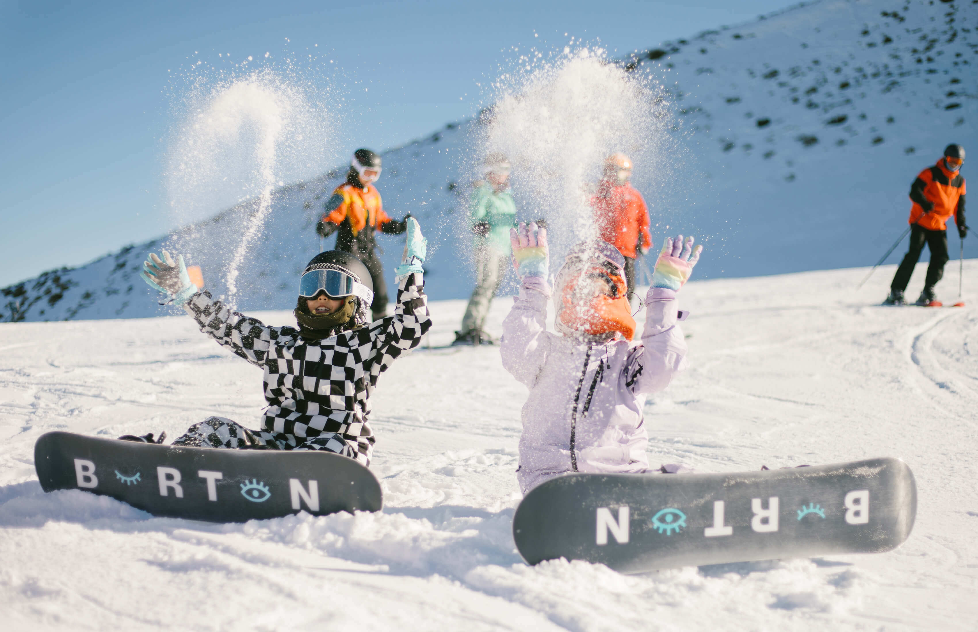 Two snowboarders sitting in snow tossing snow into the air with skiers in background on sunny day.