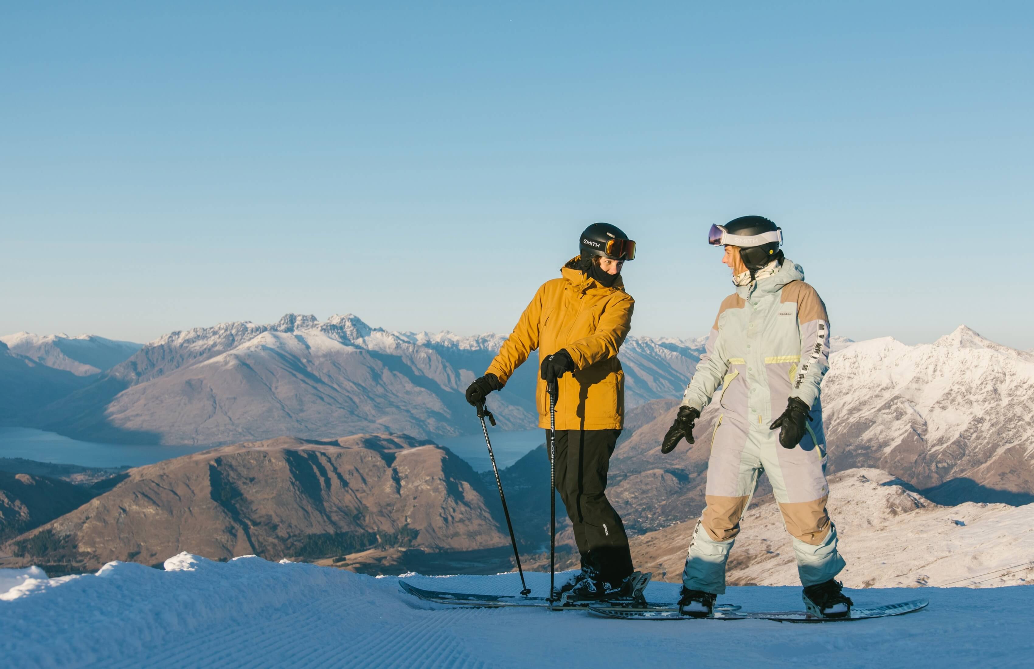 Two people in winter gear on a snowy mountain with skis and snowboard, snowy peaks behind them.