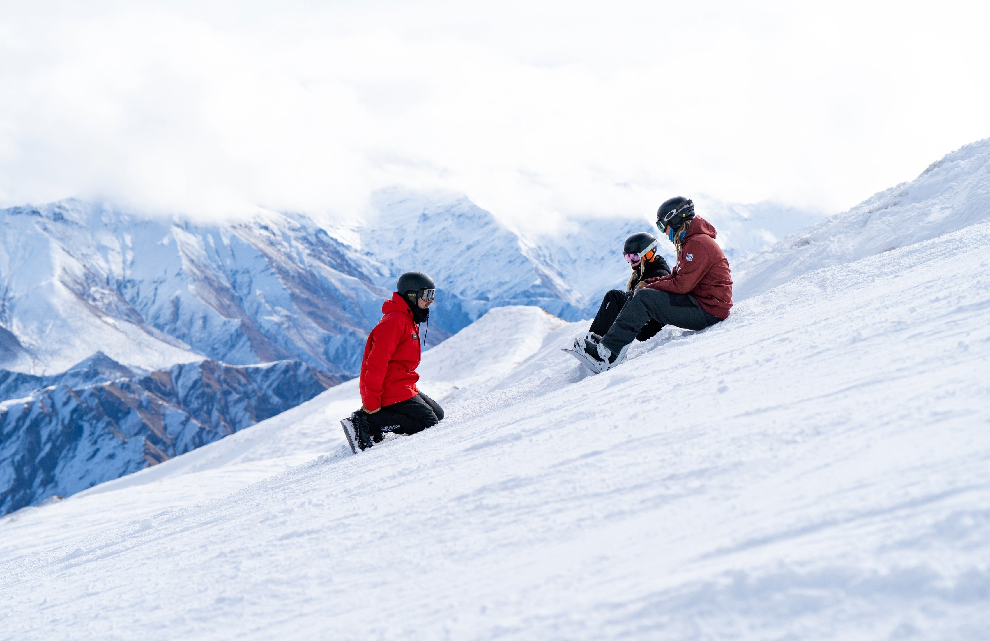 Three snowboarders resting on a snowy slope with snow-covered mountains in the background.