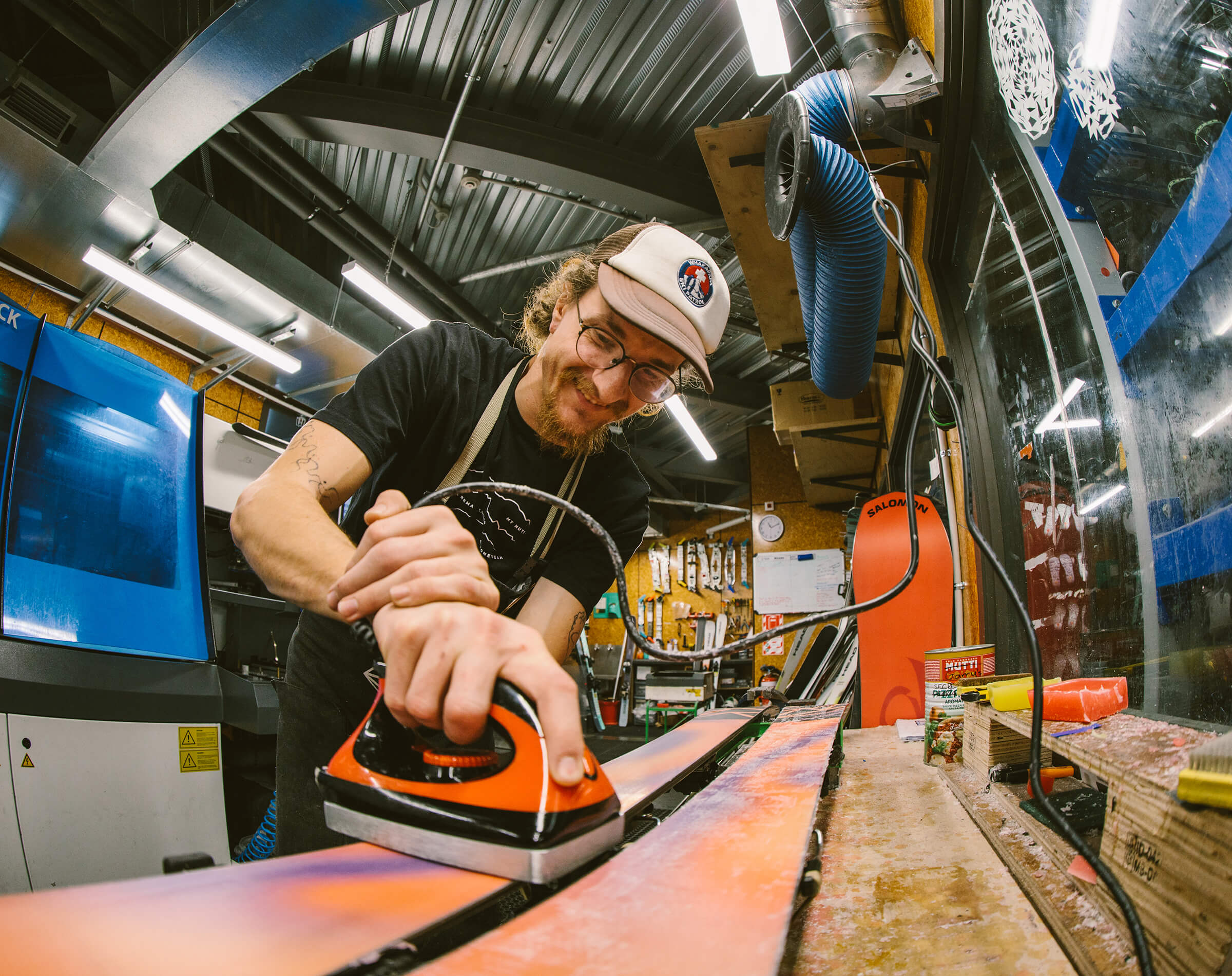 Man with glasses and cap waxing or ironing skis in a workshop filled with ski equipment.