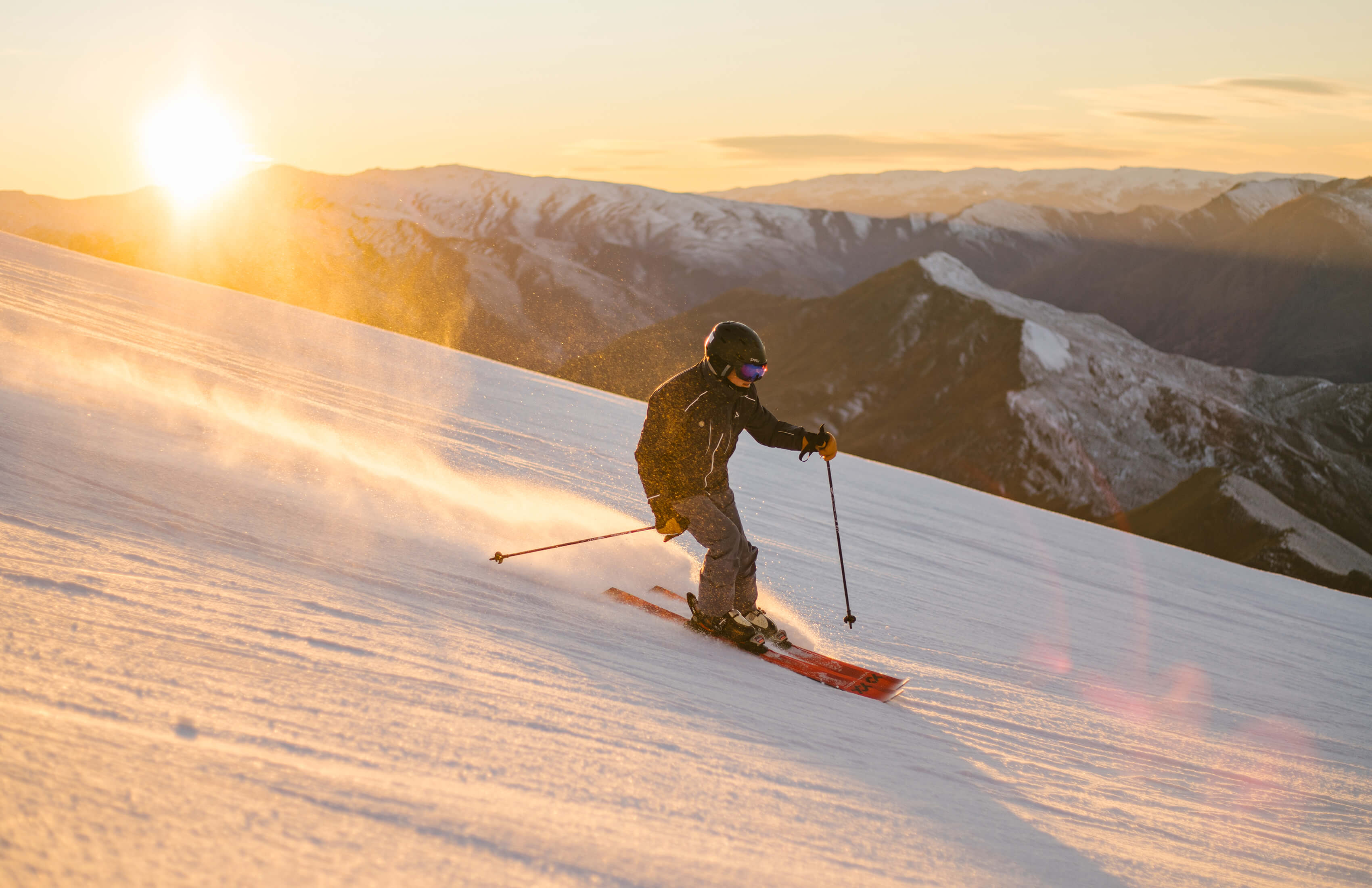 Skier in black jacket skiing down snow-covered slope at sunset with mountains in background.