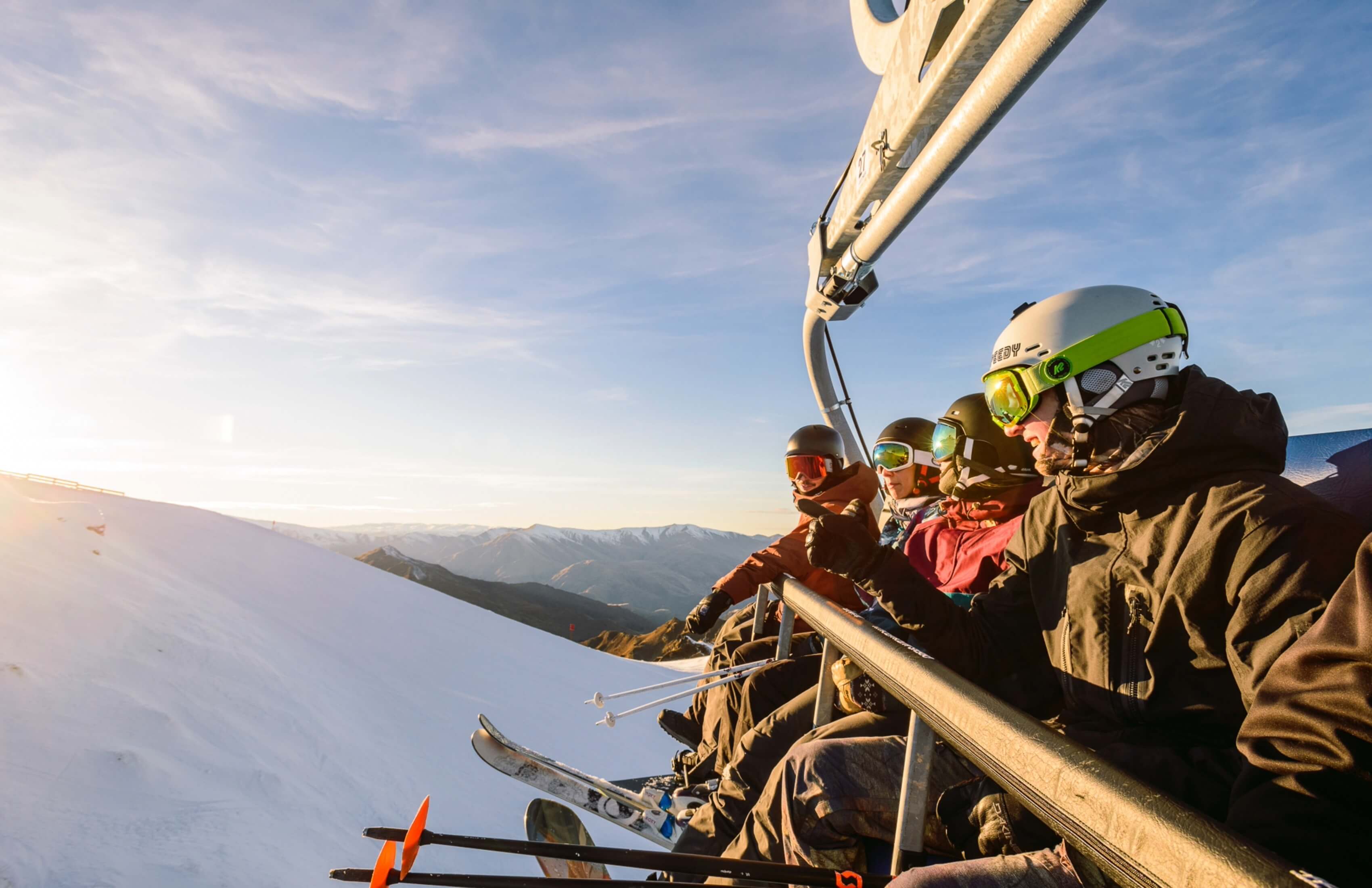 Group of skiers on a chairlift with snowy mountains and clear sky in the background at sunset.
