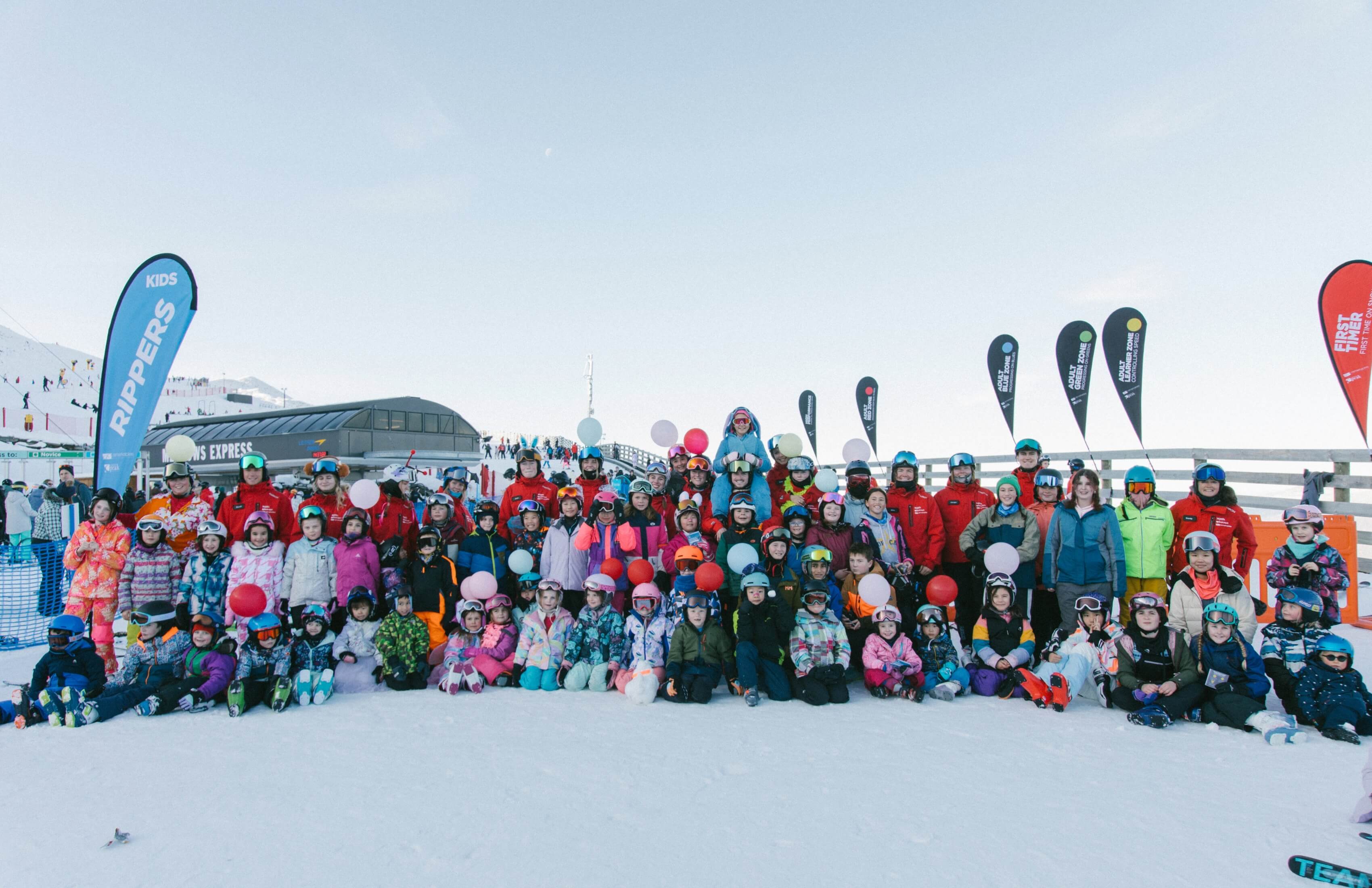 Large group of children and adults in winter gear posing on snowy ski slope with colorful balloons.