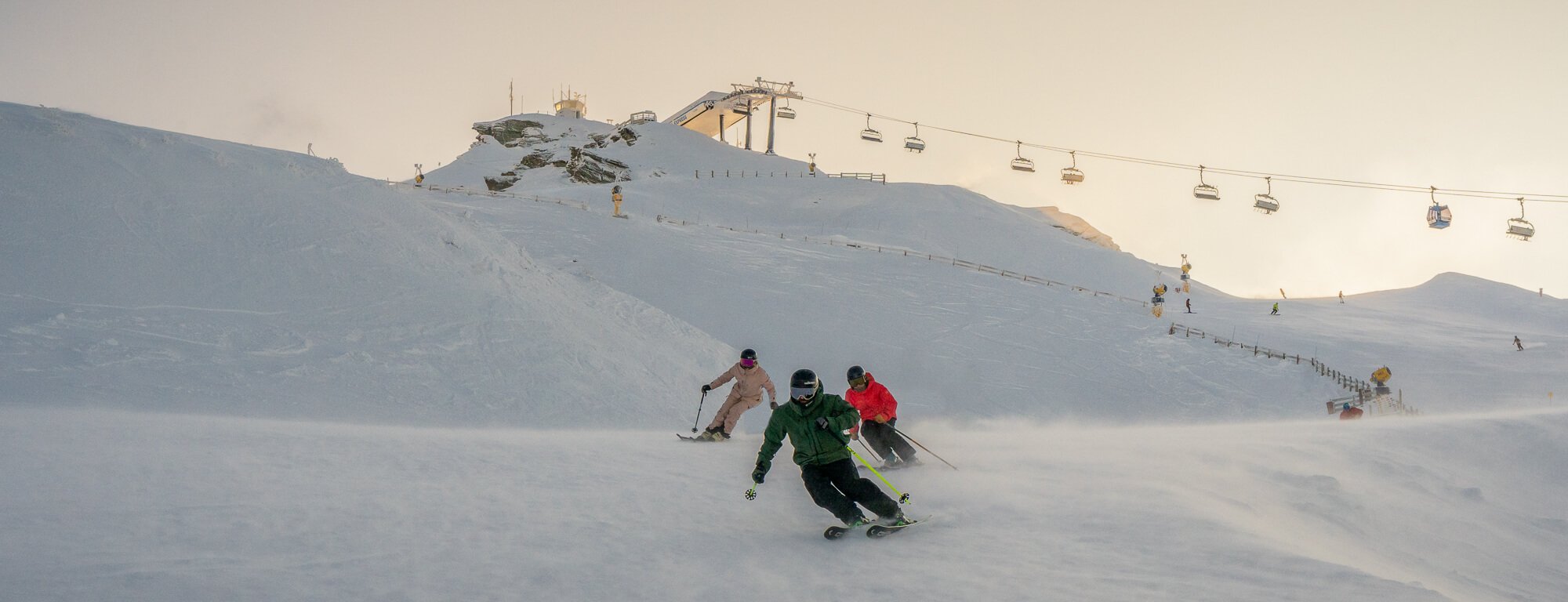 Three skiers in colorful gear skiing down a snowy mountain with ski lifts overhead.