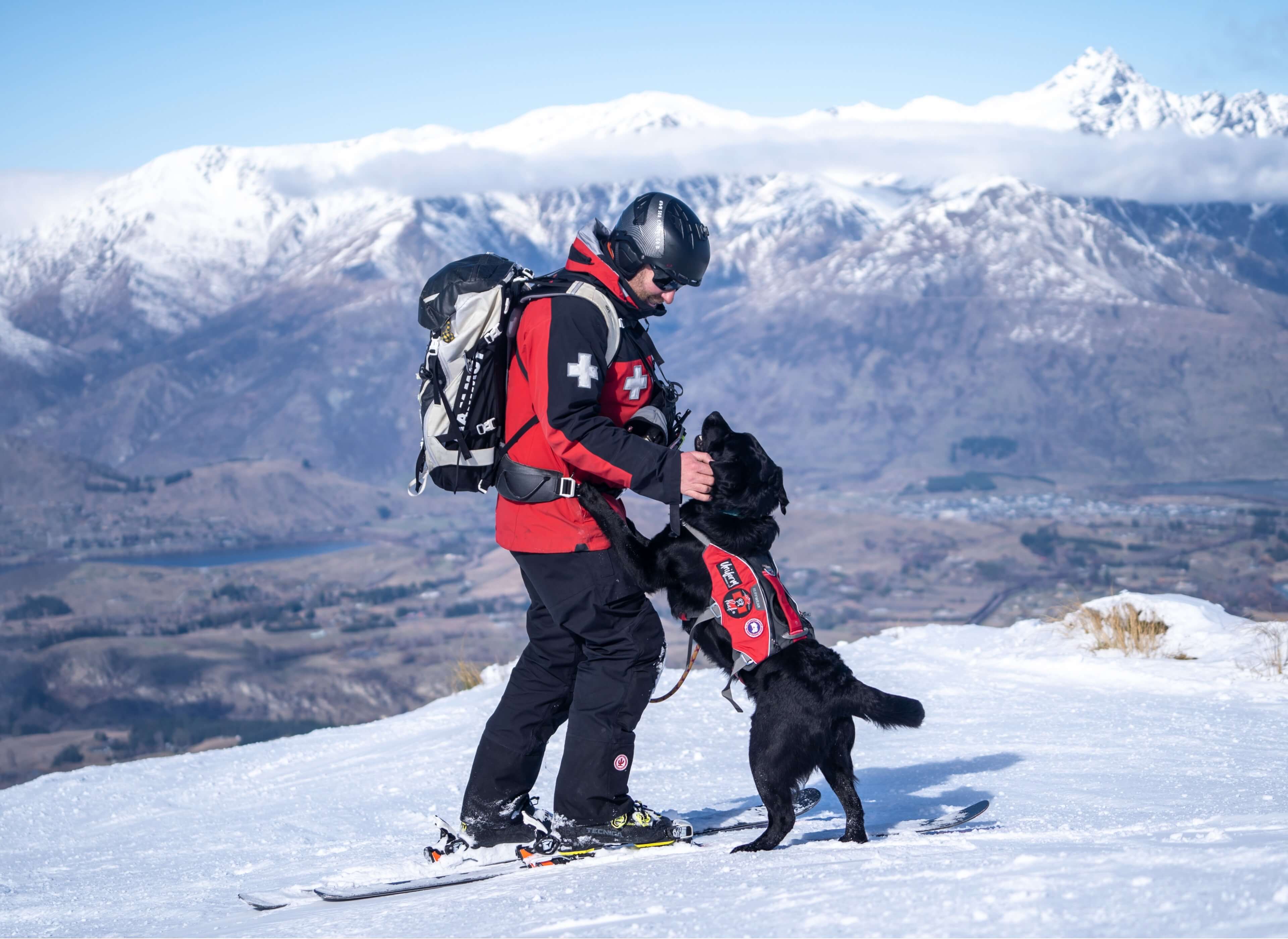 Ski patroller in red jacket with black rescue dog wearing a vest on a snowy mountain peak.