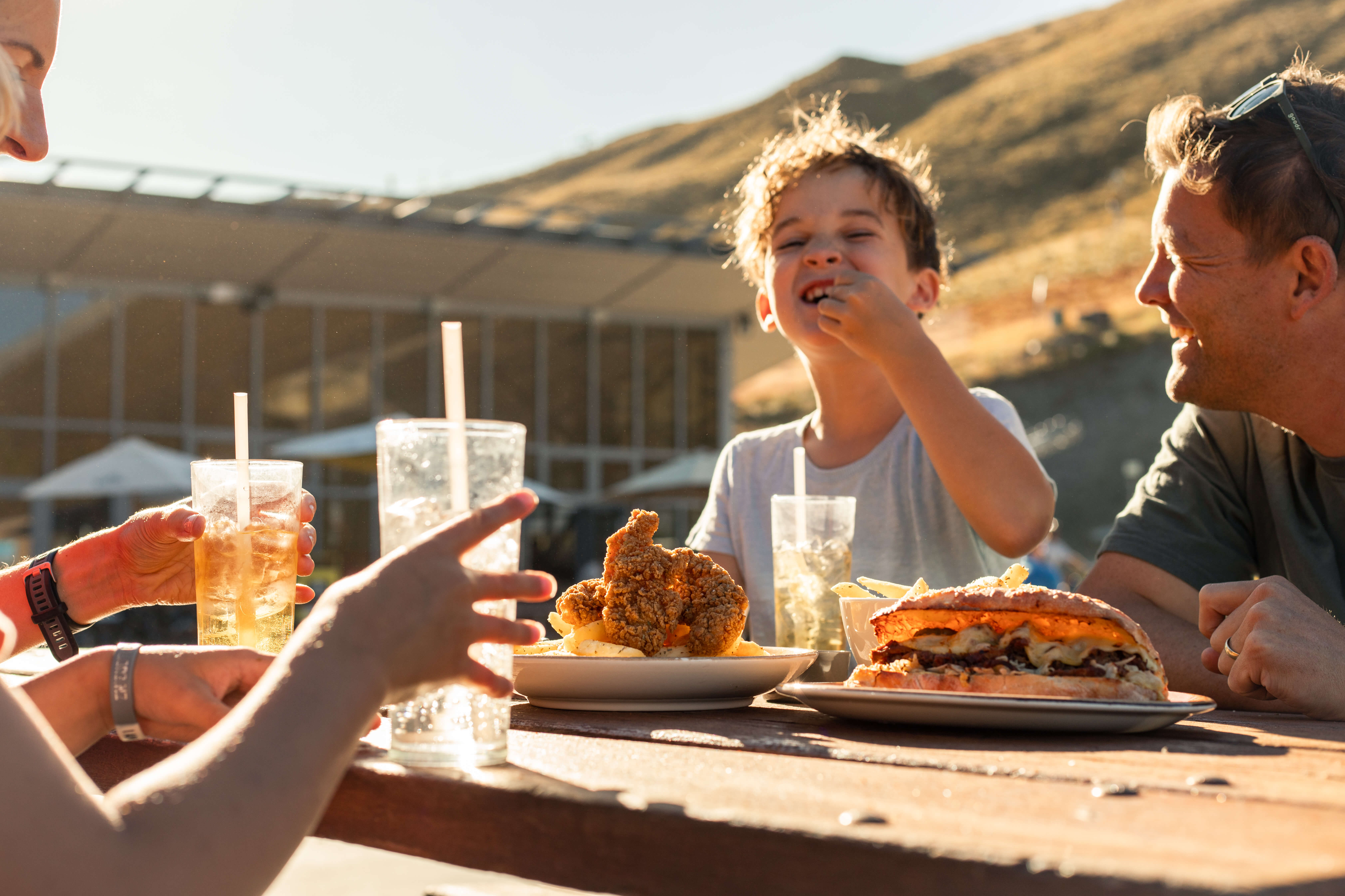 Family enjoying outdoor meal with fried chicken, sandwich, fries, and drinks on a sunny day.
