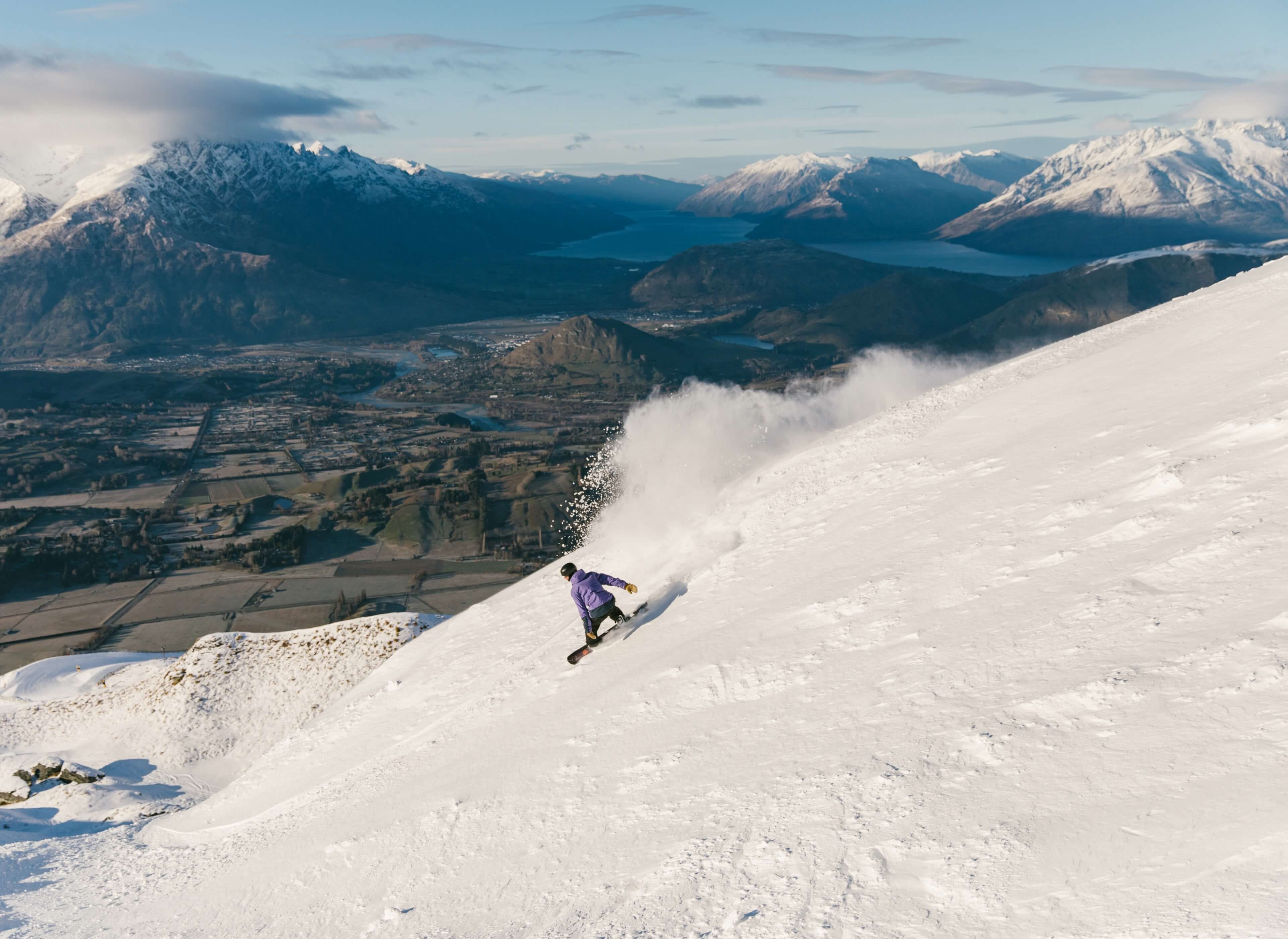 Snowboarder in purple jacket carving down a snowy mountain slope with valley and mountains in the background.