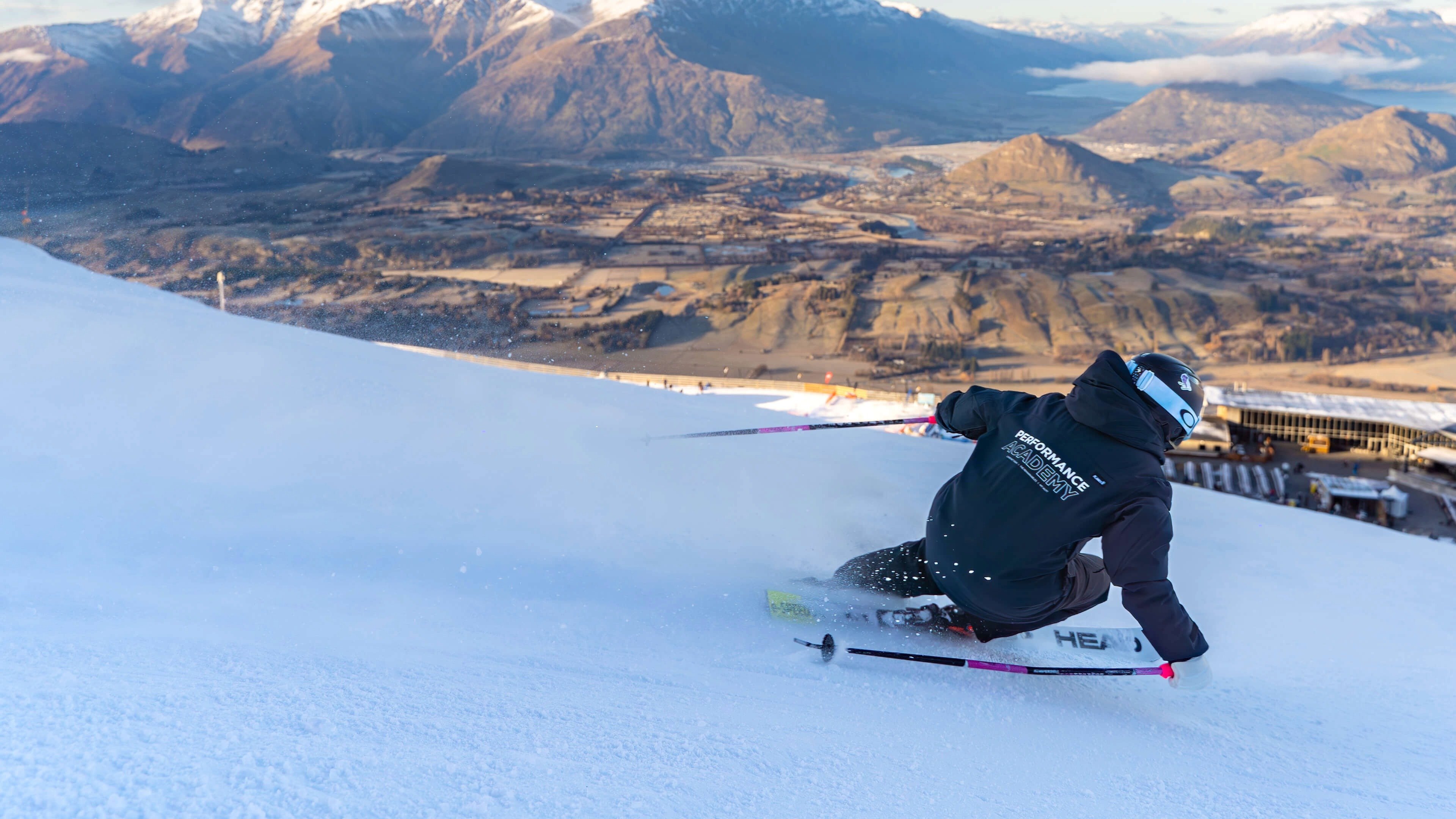 Skier in Performance Academy jacket carving a turn on a snowy slope with mountains and valley below.